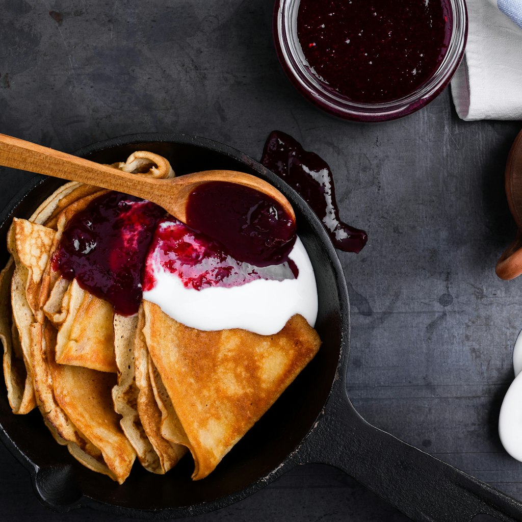Stack of pancakes in cast iron dish with sour cream and berry jam over gray background viewed from above