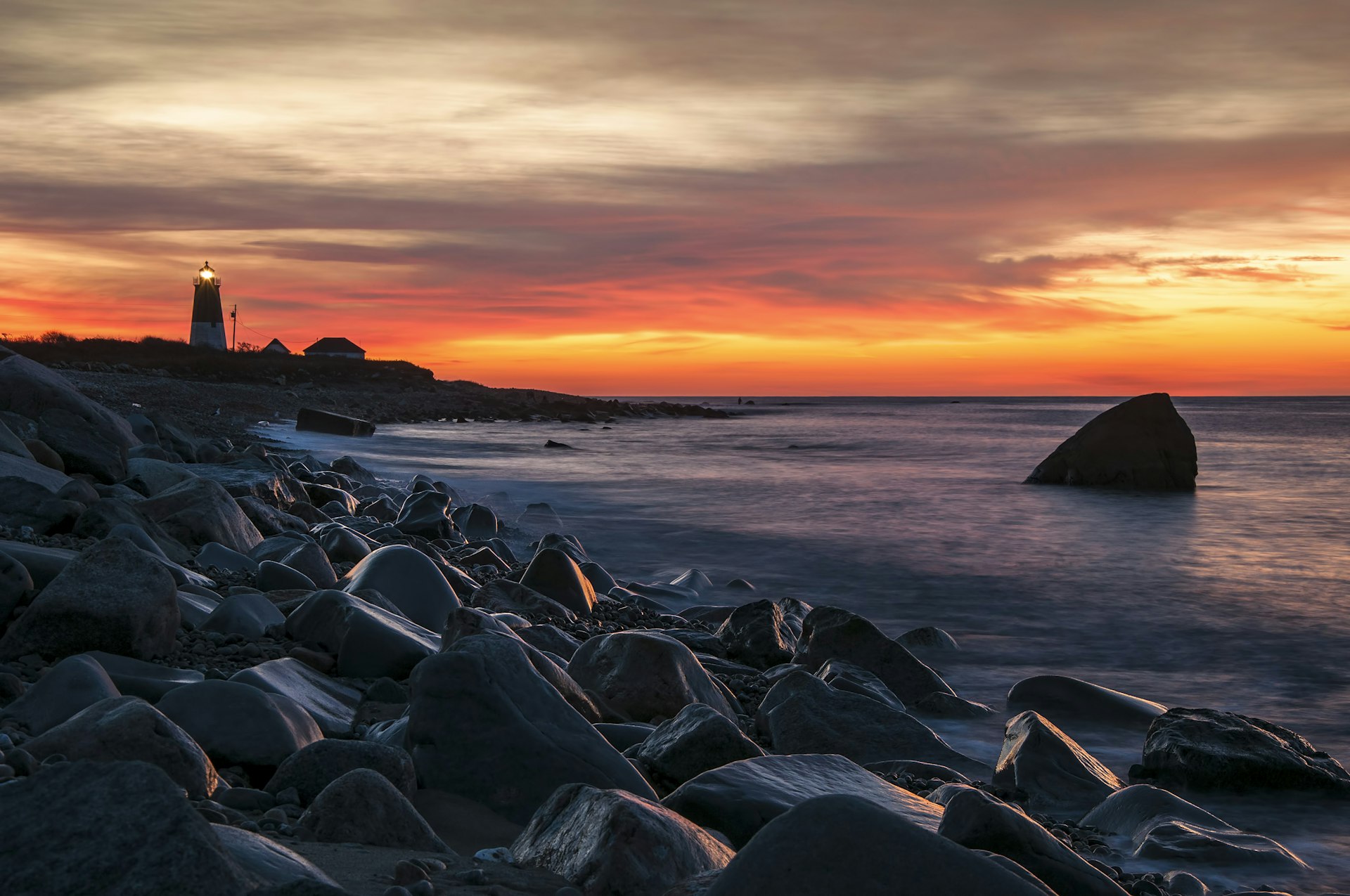 A sunrise shot on a rocky coastline, with the sky streaked with oranges and yellows. A lighthouse, to the left of the image, is silhouetted