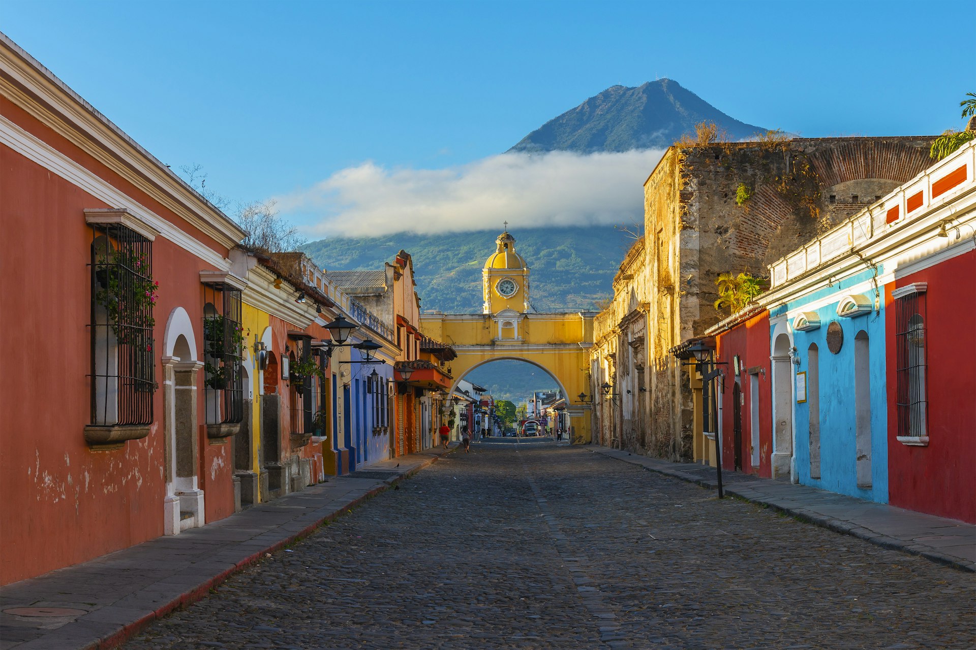 A cobbled street lined with colorful painted low-rise properties. A yellow arch with a clock on the top rises over the street. A large mountain covered with cloud looms over the town