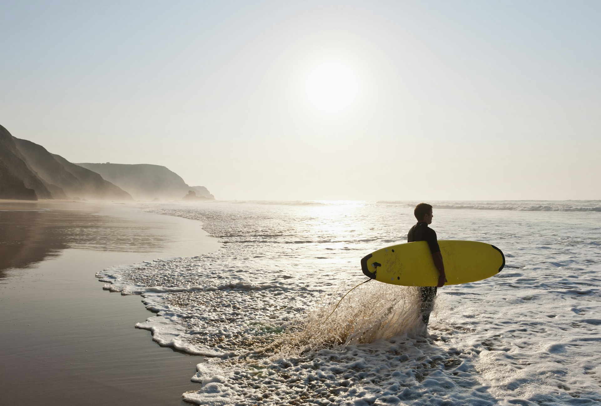 A surfer stands on a beach in morning lights. 