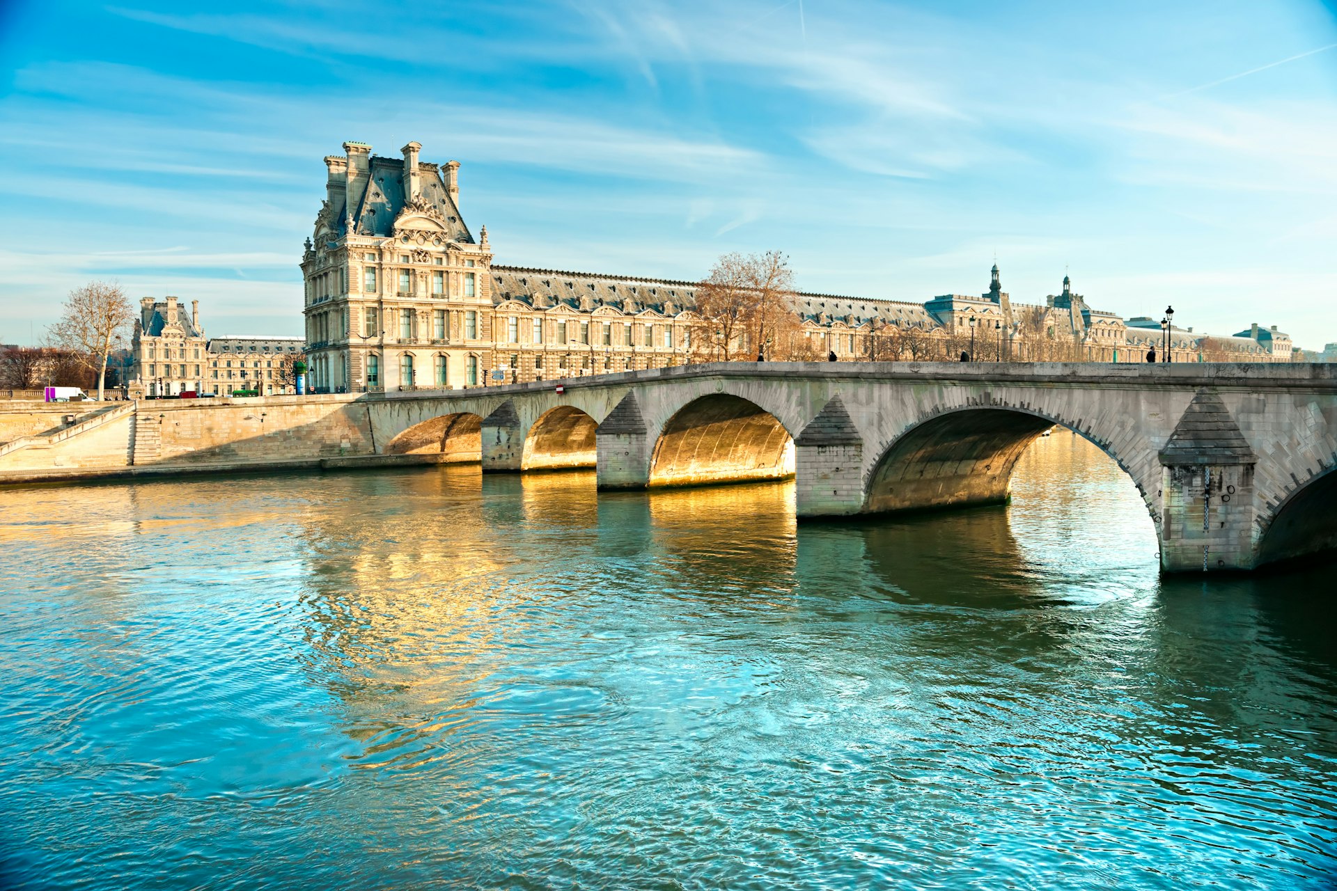 A river with an arched bridge crossing it. A classic-style large building in the background is the Louvre museum