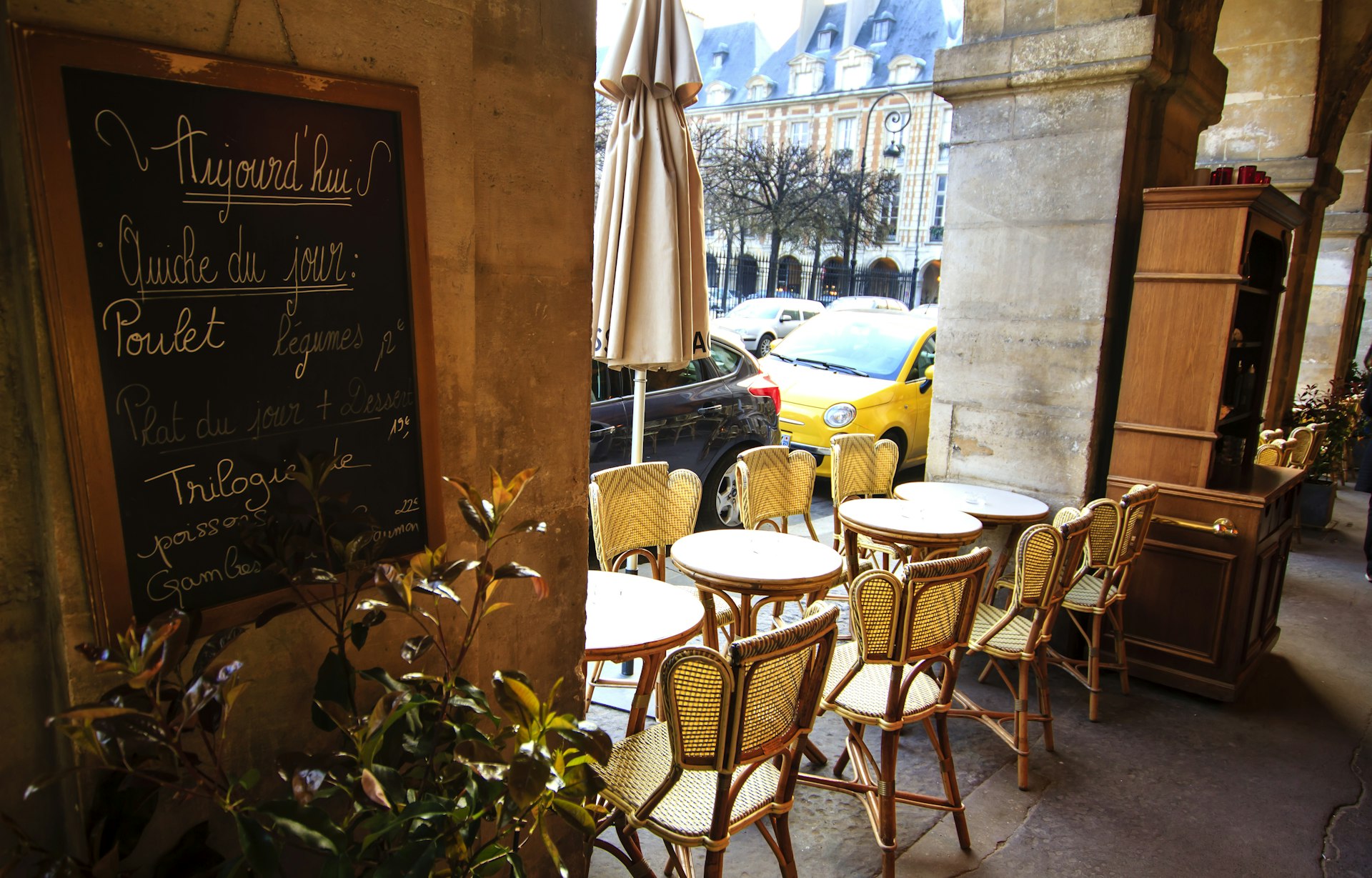 An empty pavement cafe with chairs and tables lined up between pillars. The blackboard menu is displayed on the wall