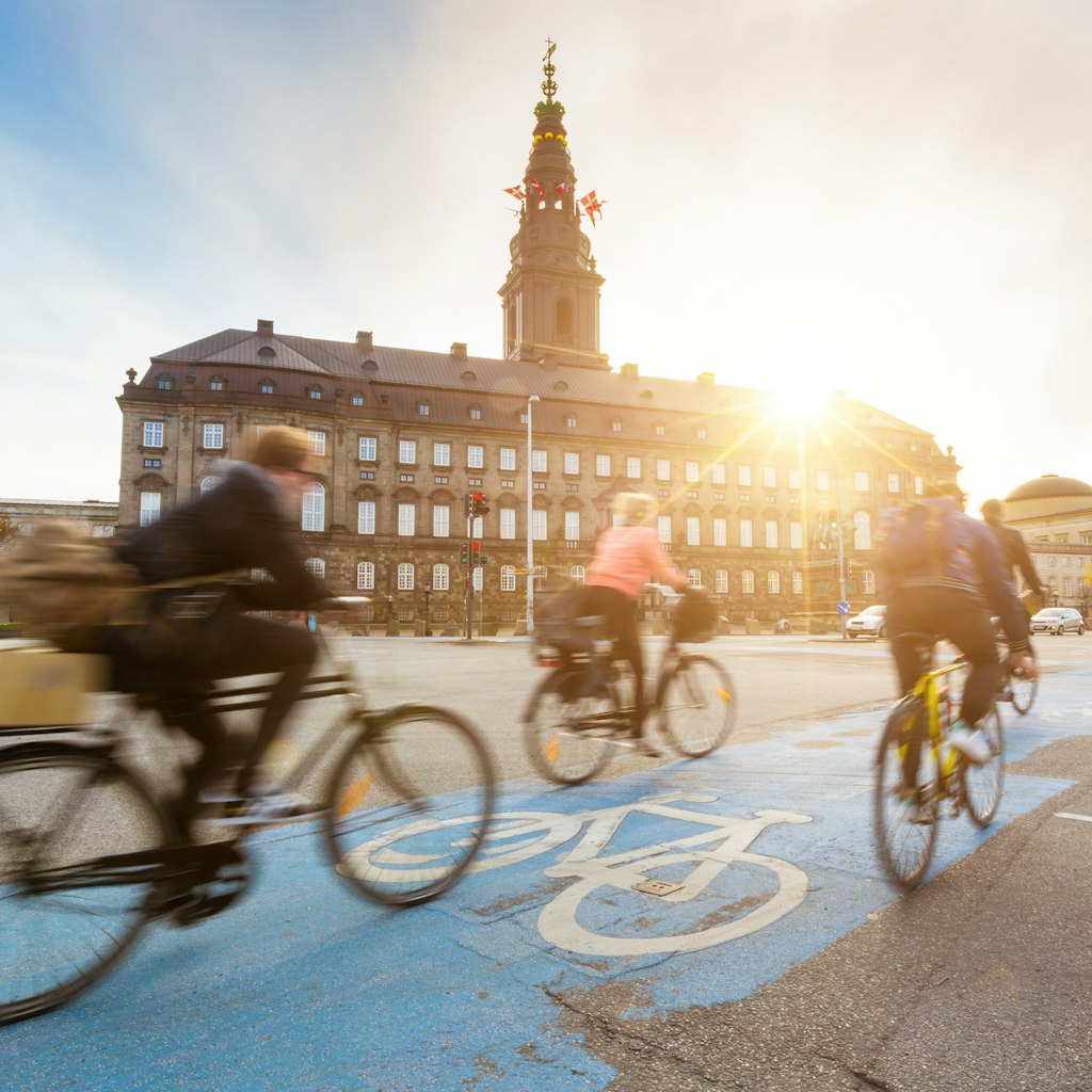 Blurred cyclists ride on a bike lane in Copenhagen, with the sun setting over Christiansborg palace.
