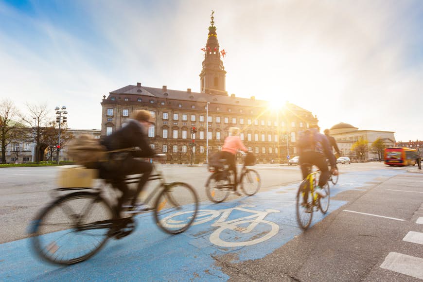 Blurred cyclists ride on a bike lane in Copenhagen, with the sun setting over Christiansborg palace