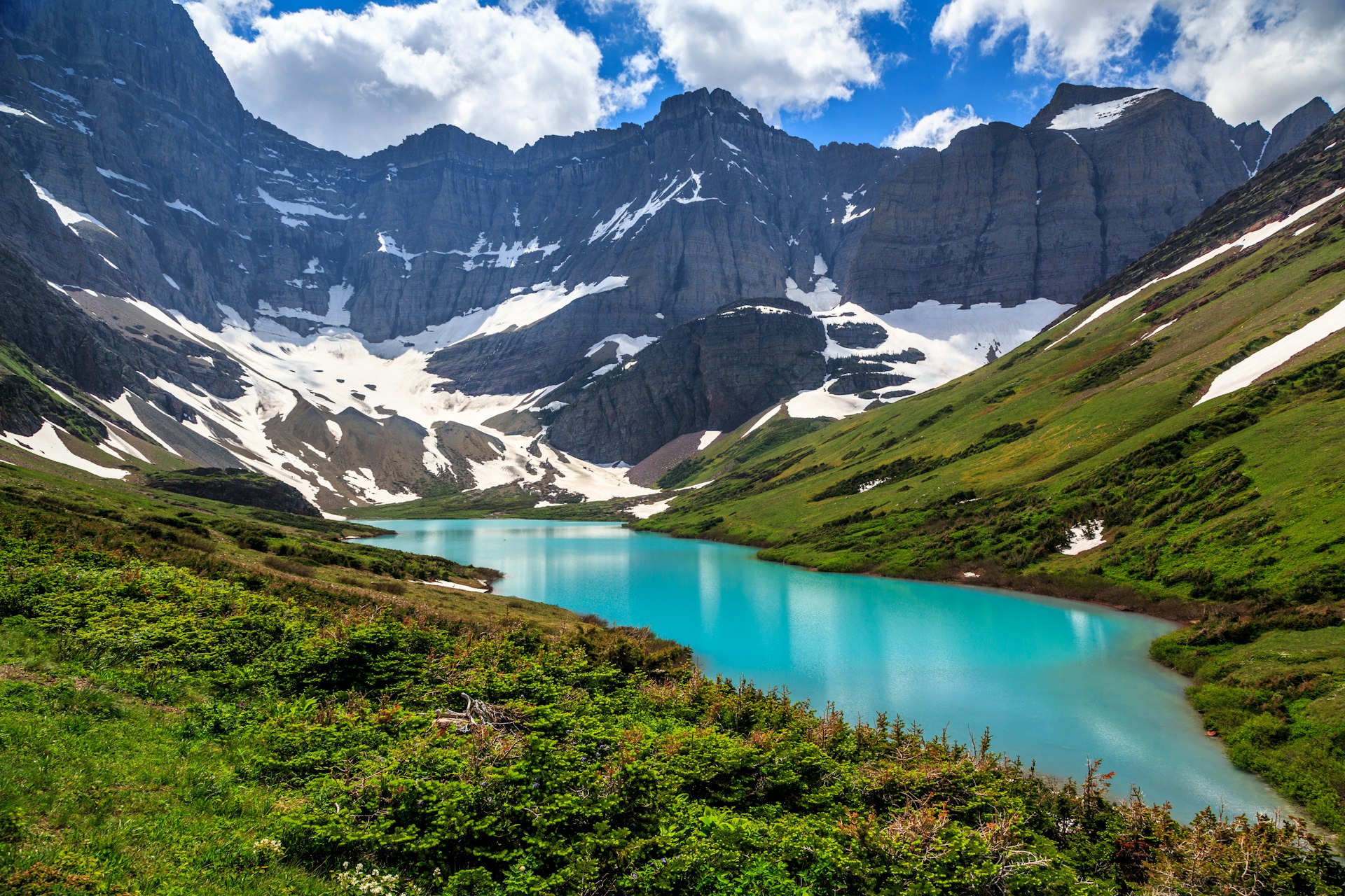 A turquoise lake surrounded by green hills and mountains with patches of snow on them