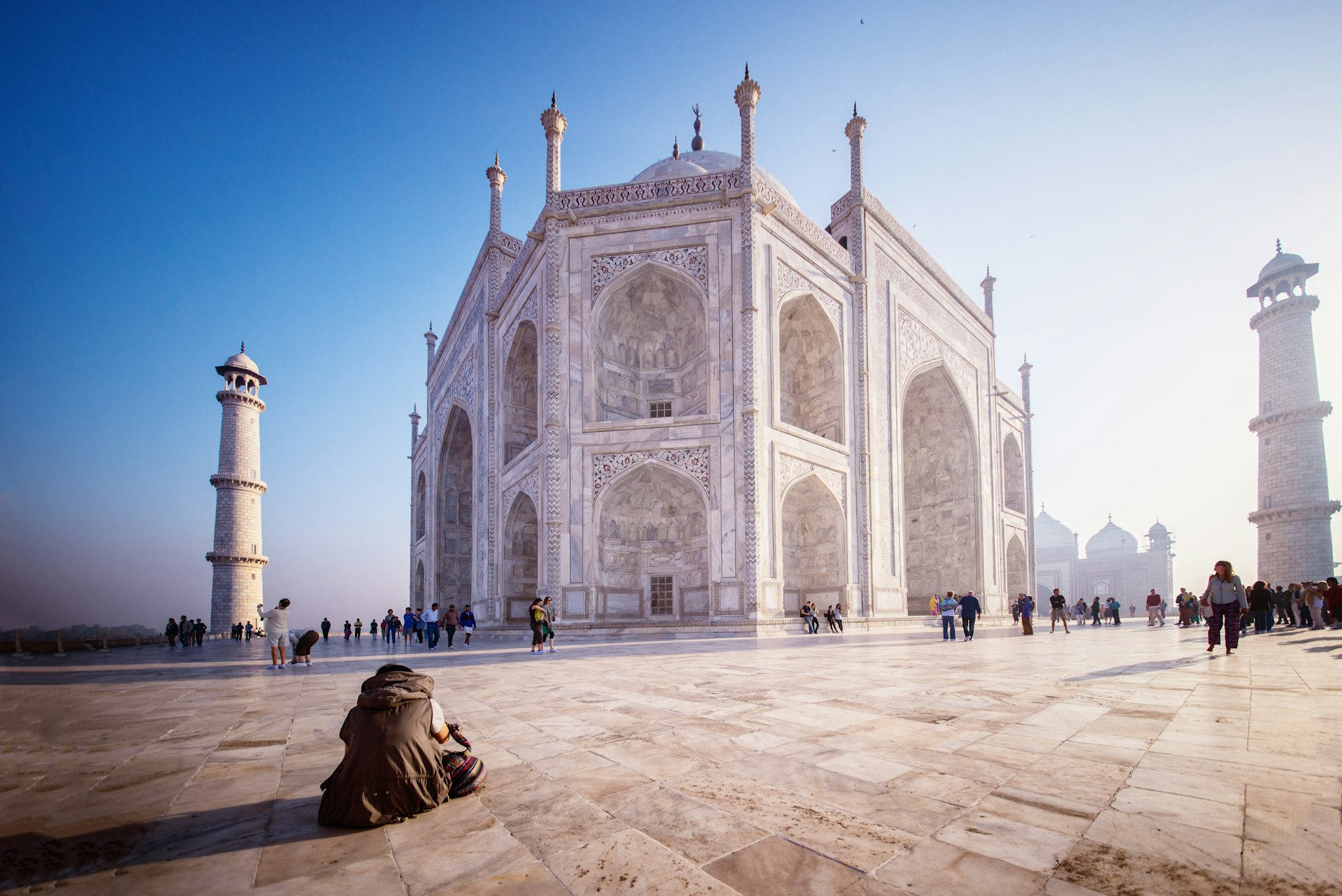 A man sits on the ground in front of the Taj Mahal in the early hours of the morning while other tourists walk around the outside of the monument.