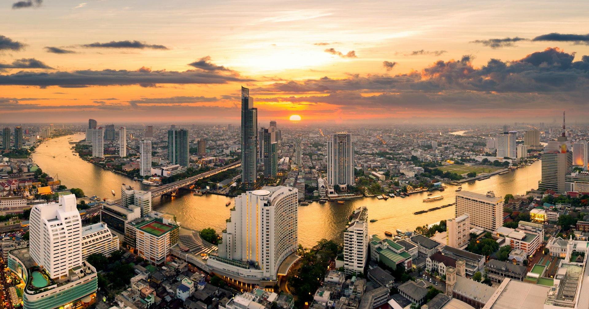 The Bangkok skyline bathed in the tangerine glow of the sun with skyscrapers reaching for the sky as a panoramic view scans around the bend of the river Chao Phraya.
