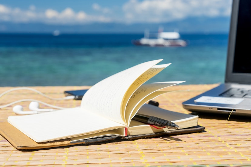 Open notepad on the table with phone and headphones, tropical sea and boat in background.