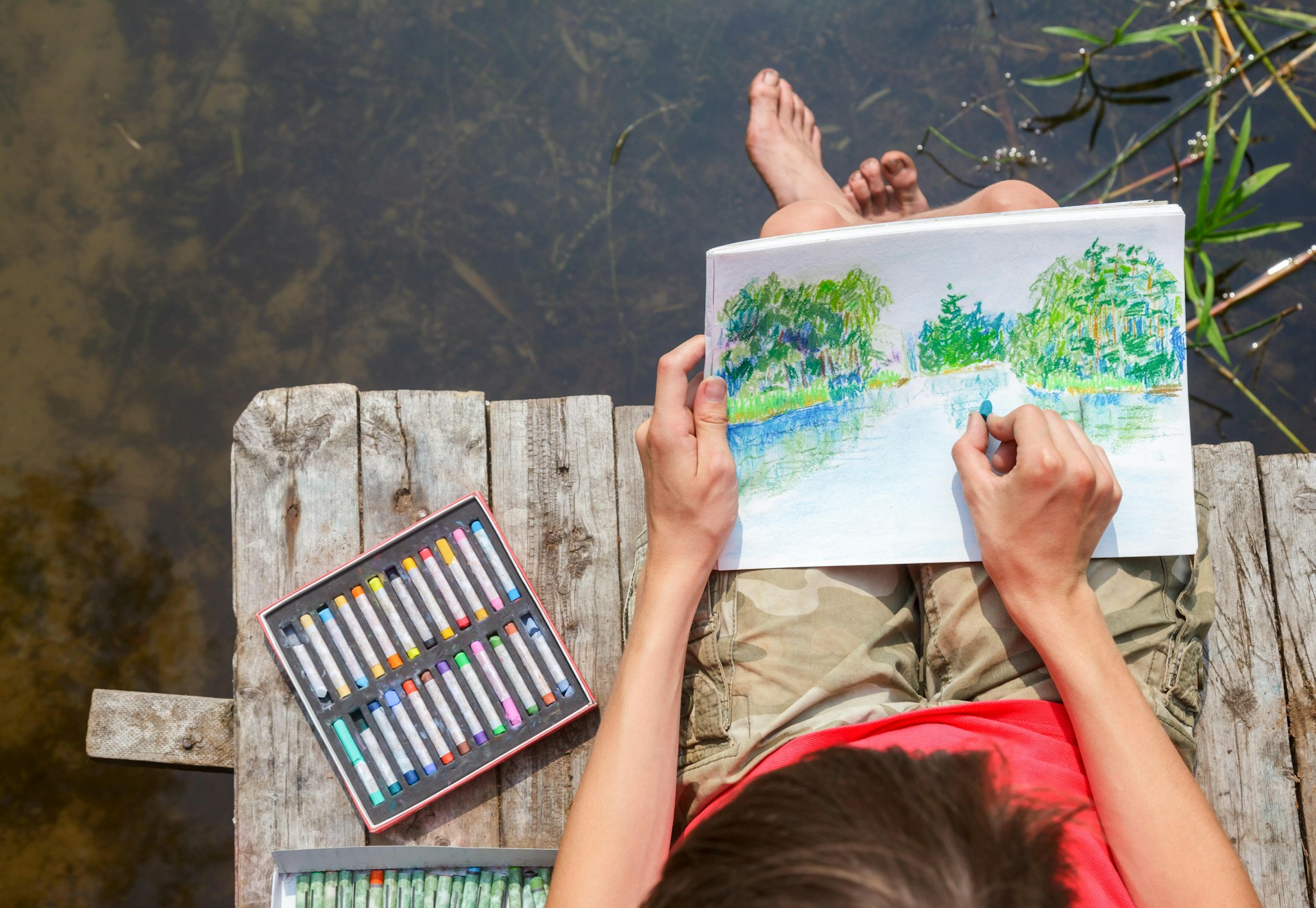 Top view of an artist sitting on a wooden dock by a forest lake drawing with pastels