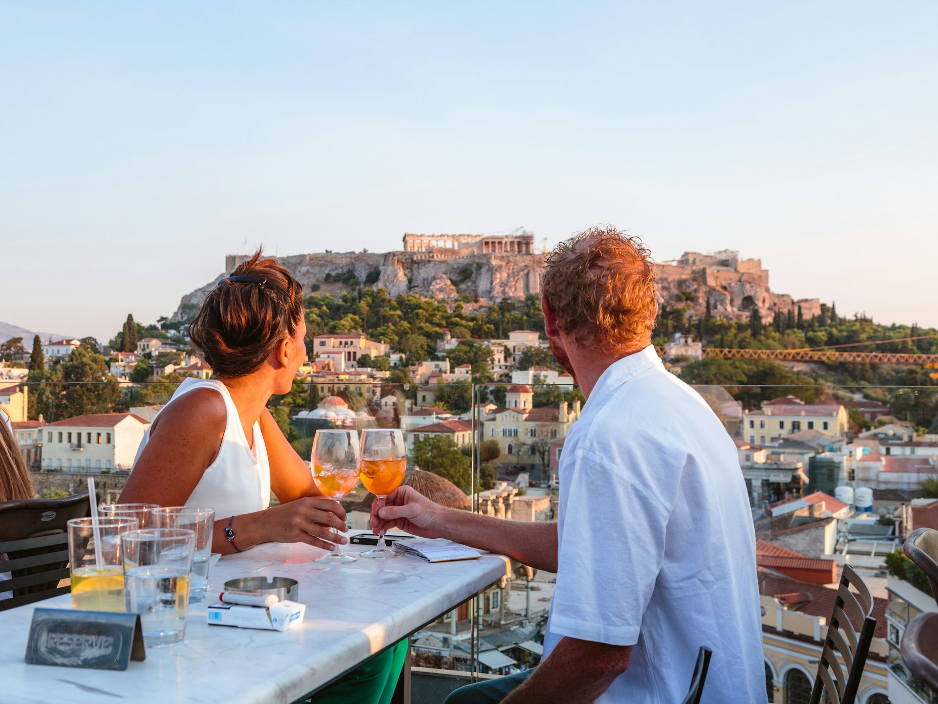 A man and a woman drinking wine and having food across the valley from the Acropolis at sunset in Athens, Greece.
