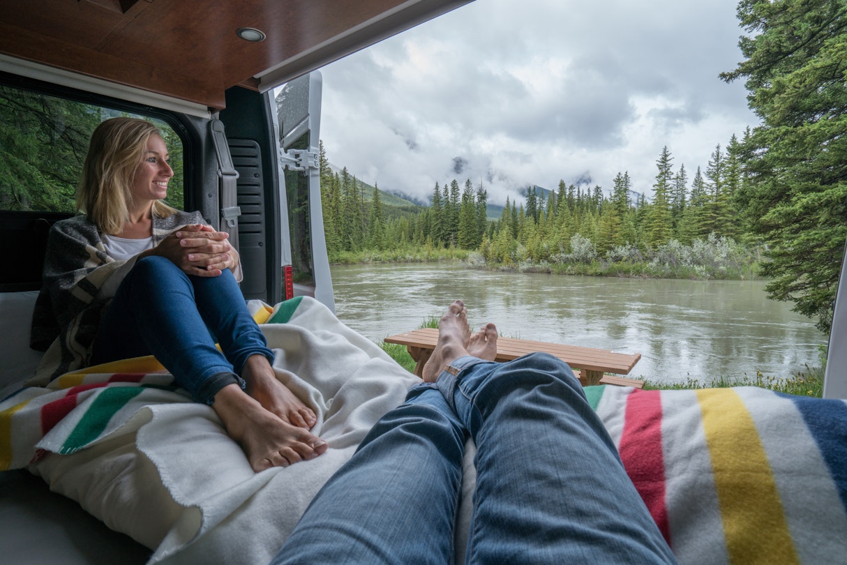 Young couple relaxing in the back of their camper van near a river and cloud-covered mountains in Alberta.