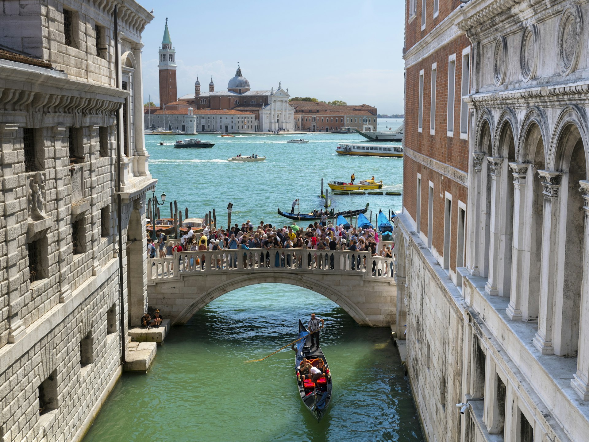 A shot down a narrow canal towards a bridge that is packed with people. A gondola passes underneath