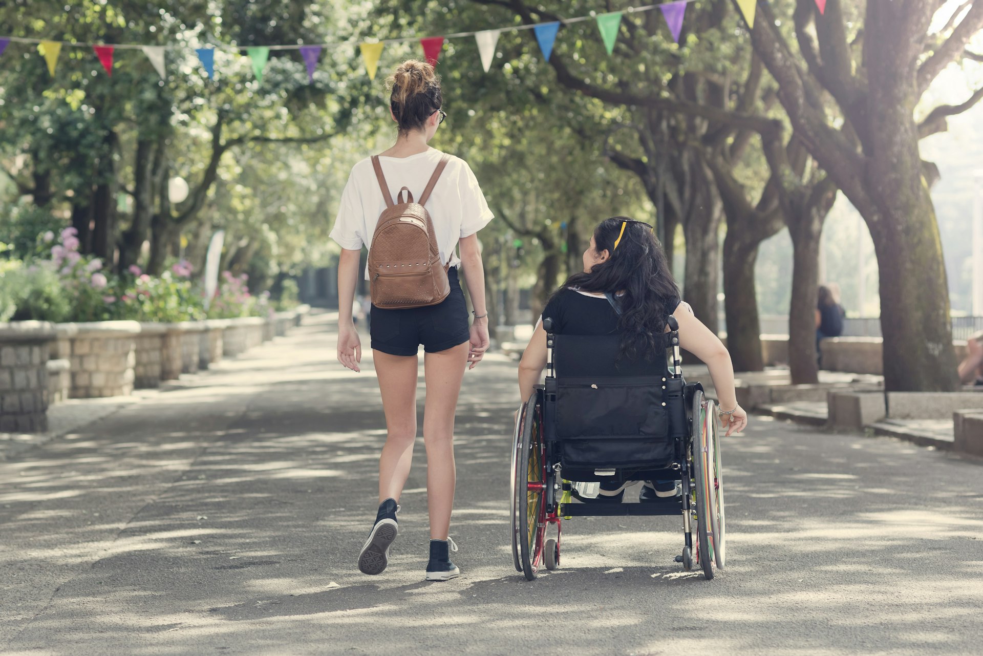 Young woman walks alongside her friend in a wheelchair in the Italian town of Tempio Pausania. The walkway is wide and flanked by greenery.