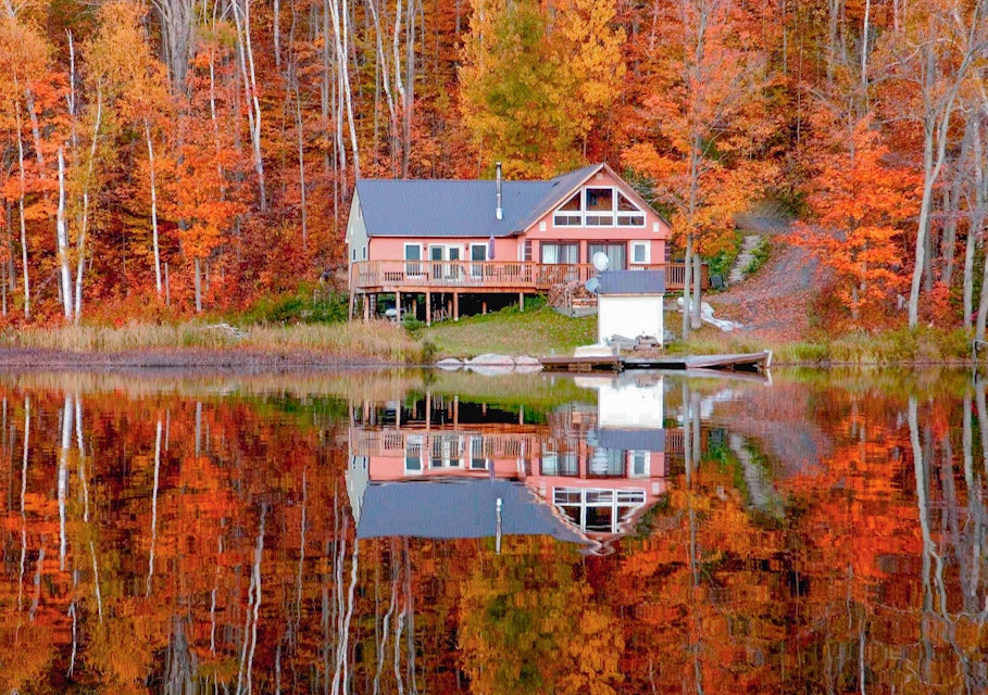 Cabin in Ontario surrounded by autumn trees and reflected in the still water of a lake.