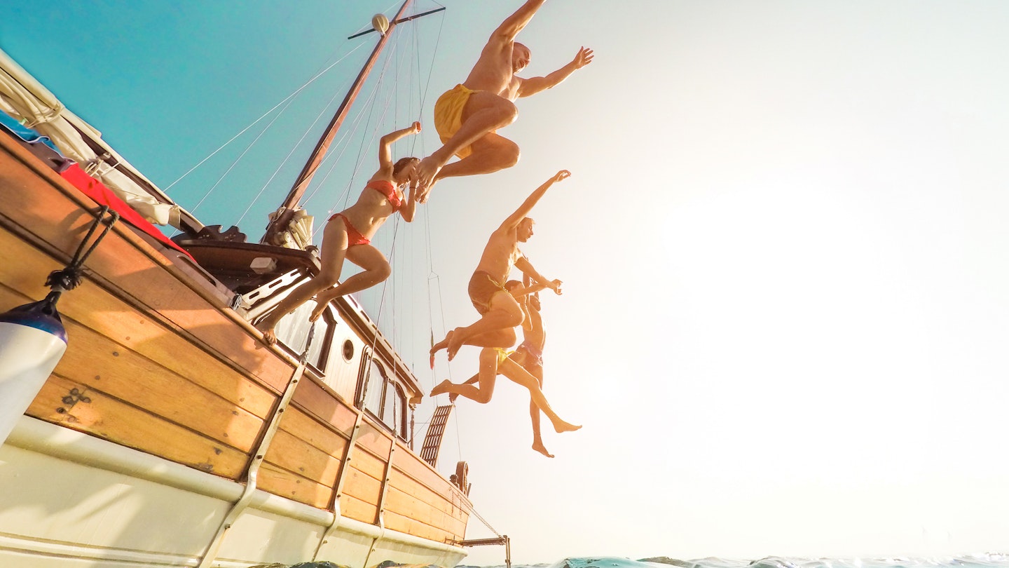 Five people jumping from a sailboat into the ocean during summer.