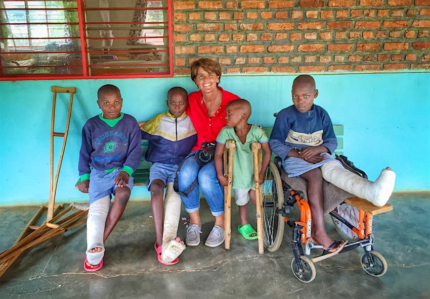 A woman sits on a bench with a group of four children. Two children have a leg in a cast. One is resting on crutches. The fourth child is in a wheelchair