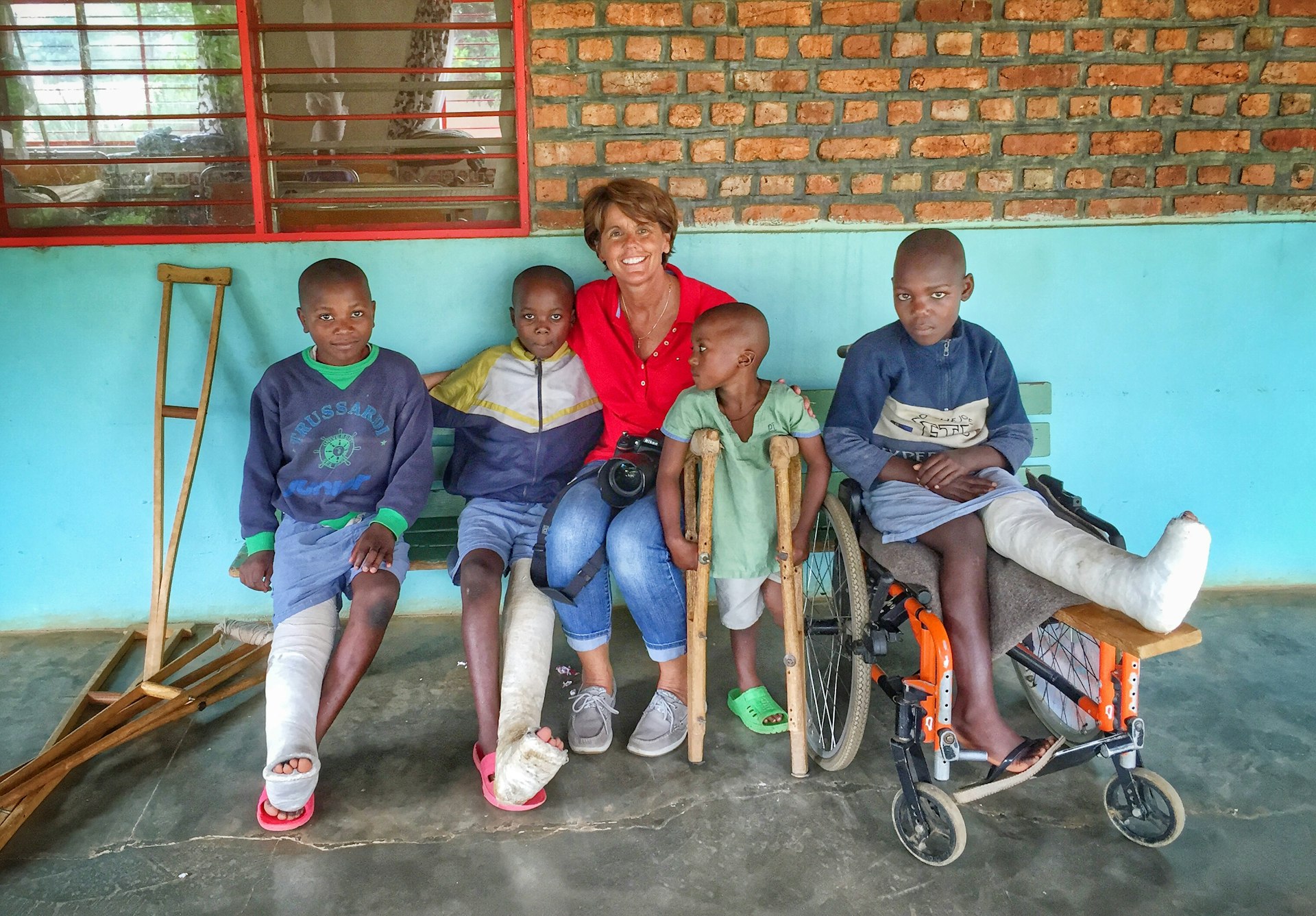 A woman sits on a bench with a group of four children. Two children have a leg in a cast. One is resting on crutches. The fourth child is in a wheelchair