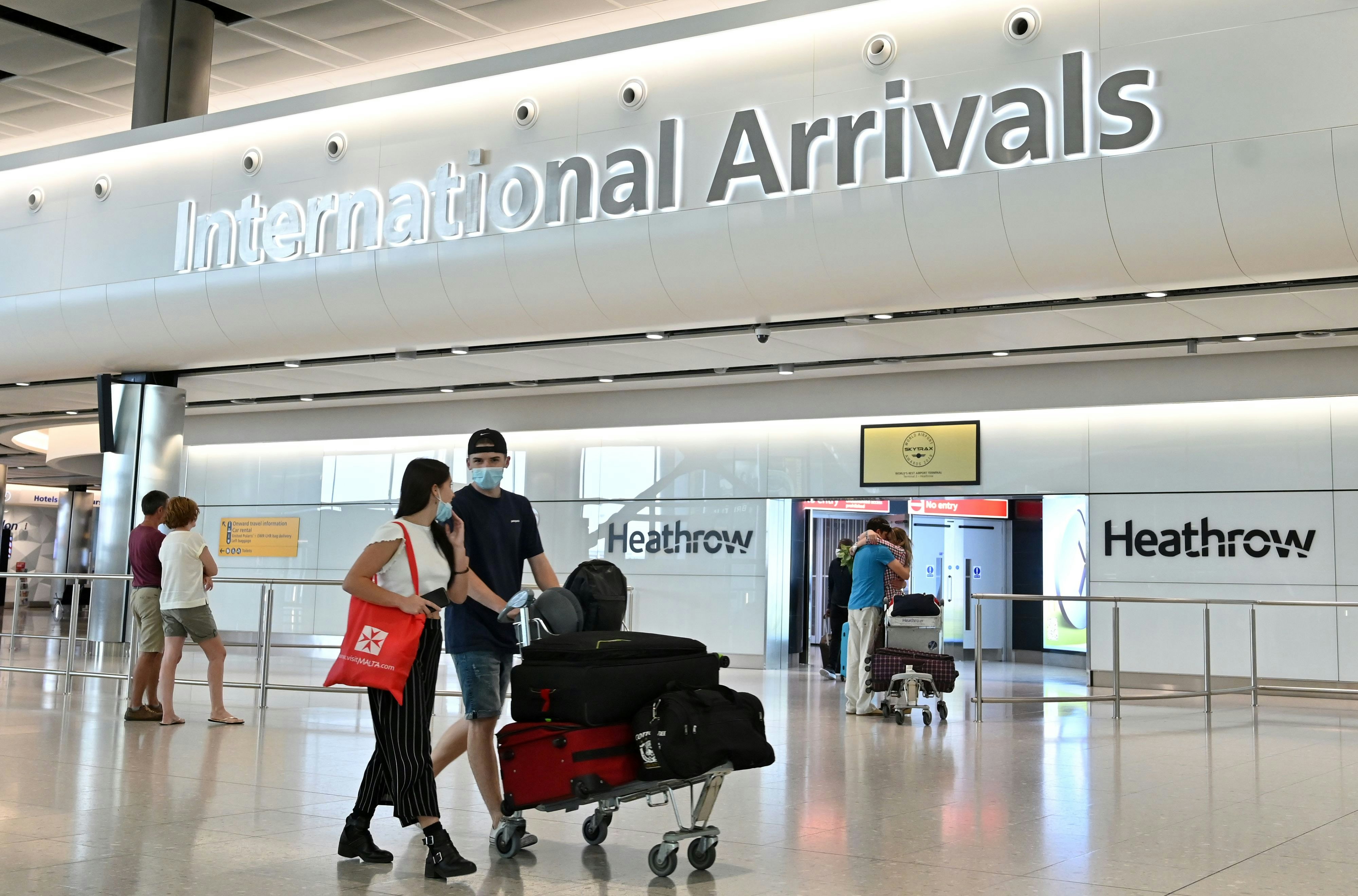 A couple wearing face masks push their luggage on a trolley in front of the International Arrivals sign at London Heathrow Airport