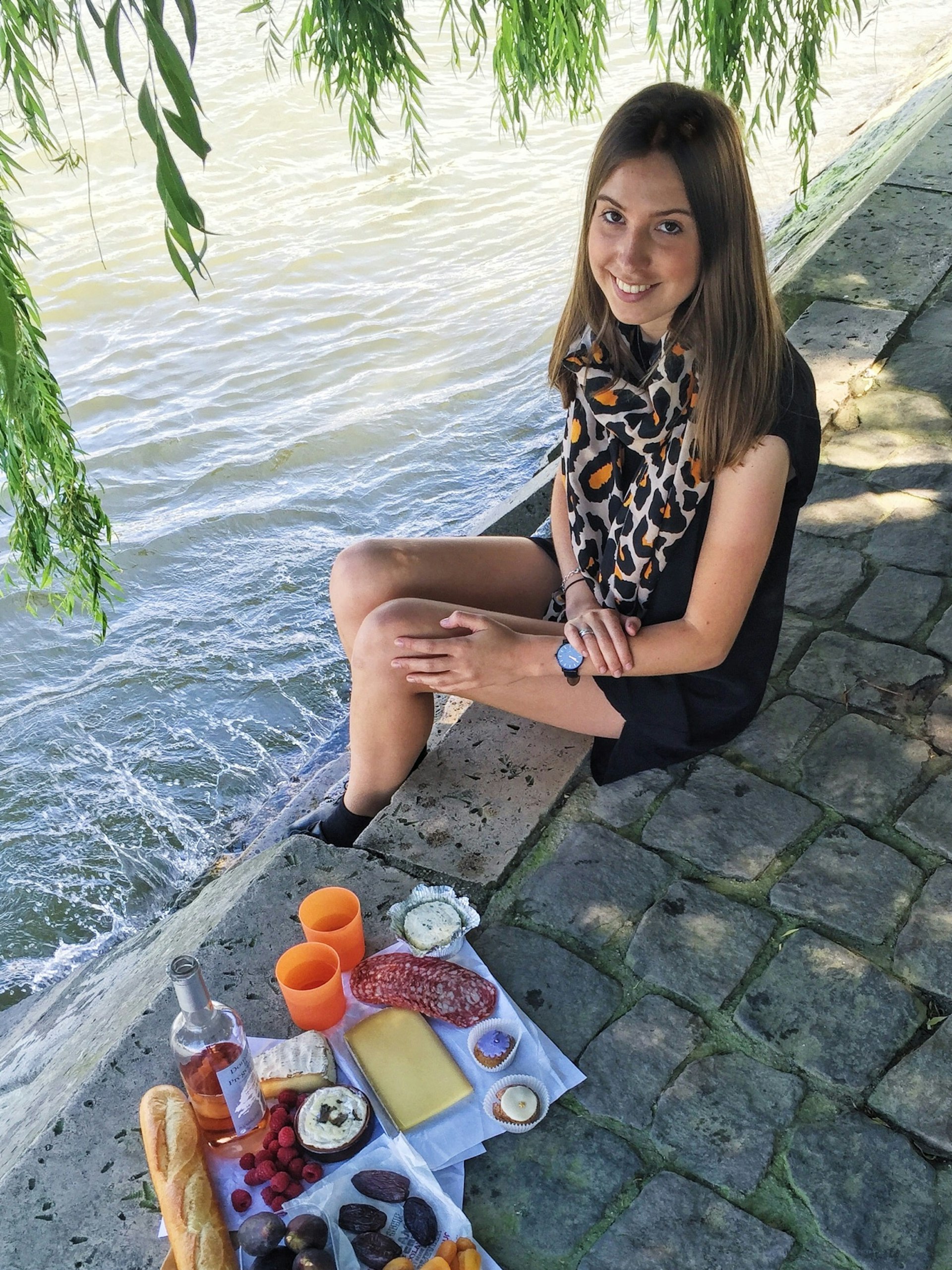 A woman smiles at the camera. She sits on the bank of a river with a small picnic laid out next to her