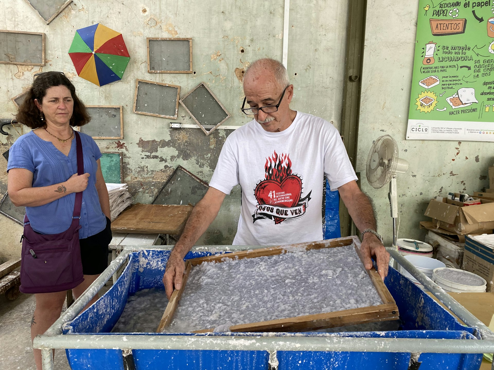 A man holding a mesh frame stands at a vat of pulp demonstrating the paper-making process to a nearby tourist