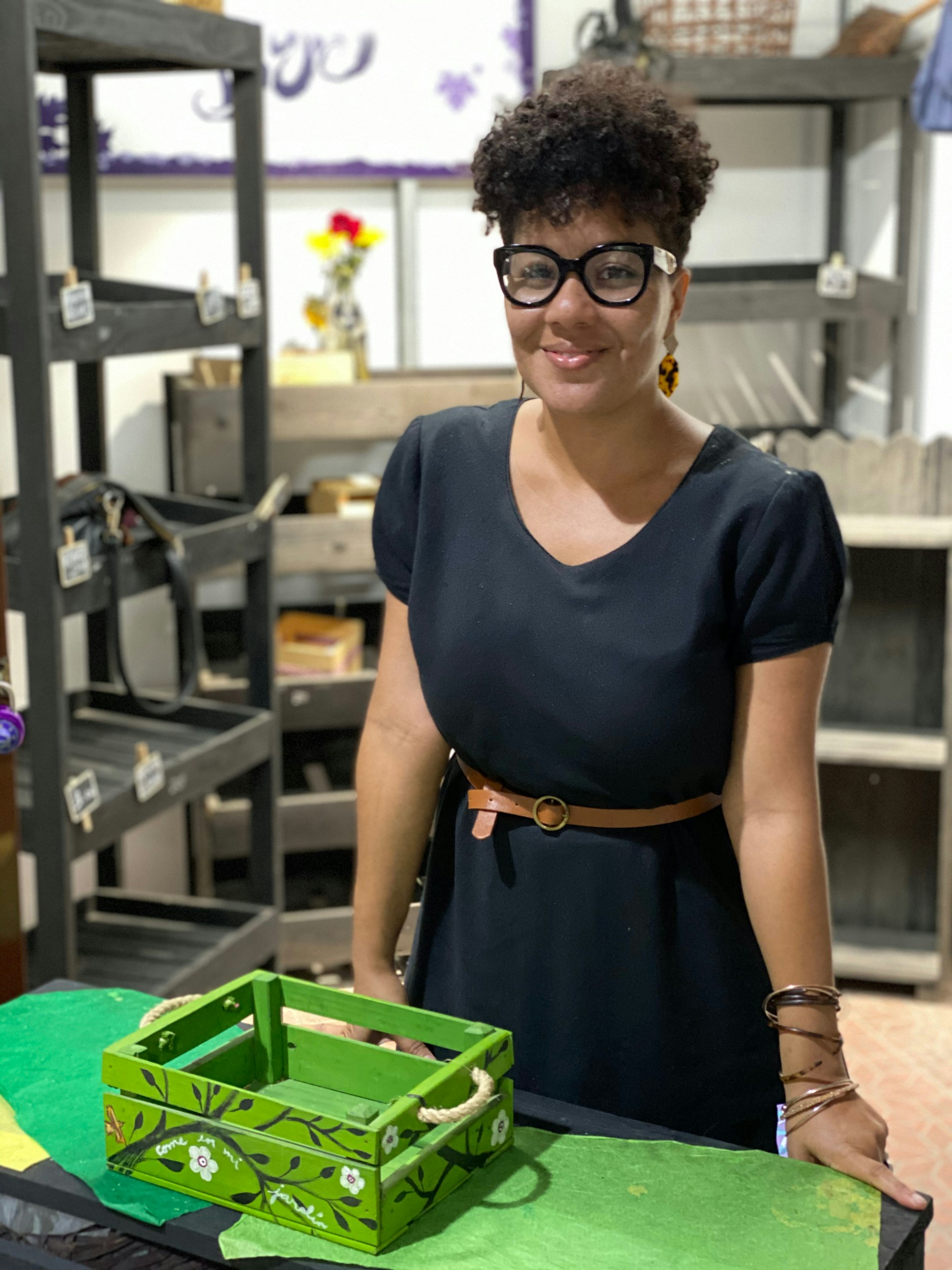 A woman wearing a black dress and glasses smiles directly at the camera. She's standing at a table with an empty green wooden crate on it