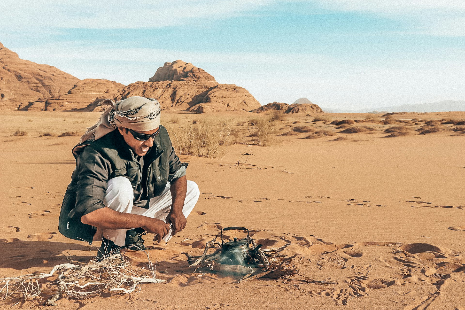 A man squats in desert sand, tending to a fire