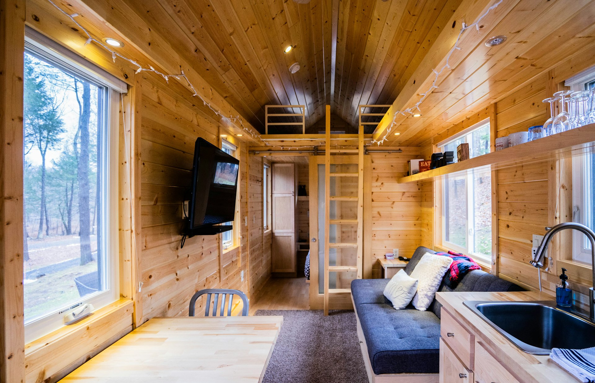 The interior of a narrow wooden cabin, with a kitchen sink, dining table, seating area and wooden ladder leading up to a loft bunk space.