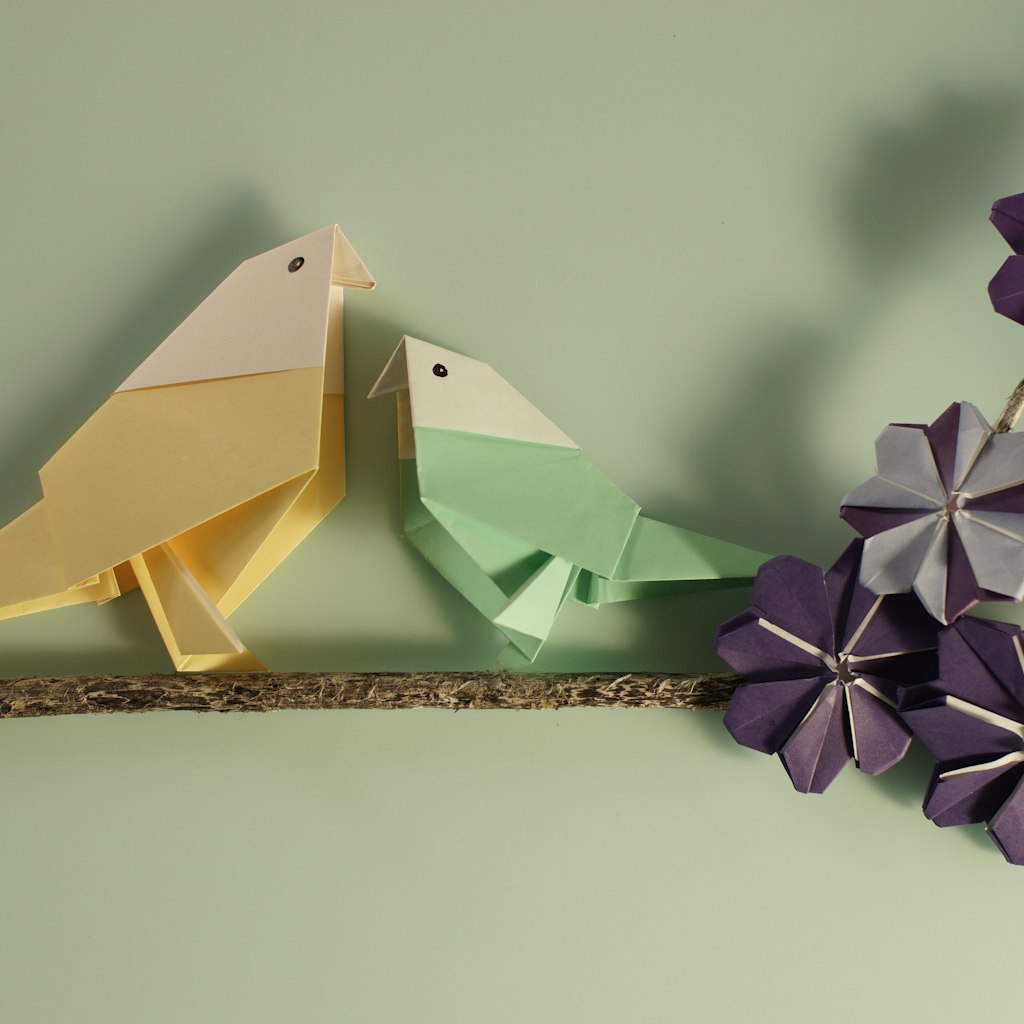 To paper origami birds perched on a tree branch with purple cosmos origami flowers on. Space for copy.