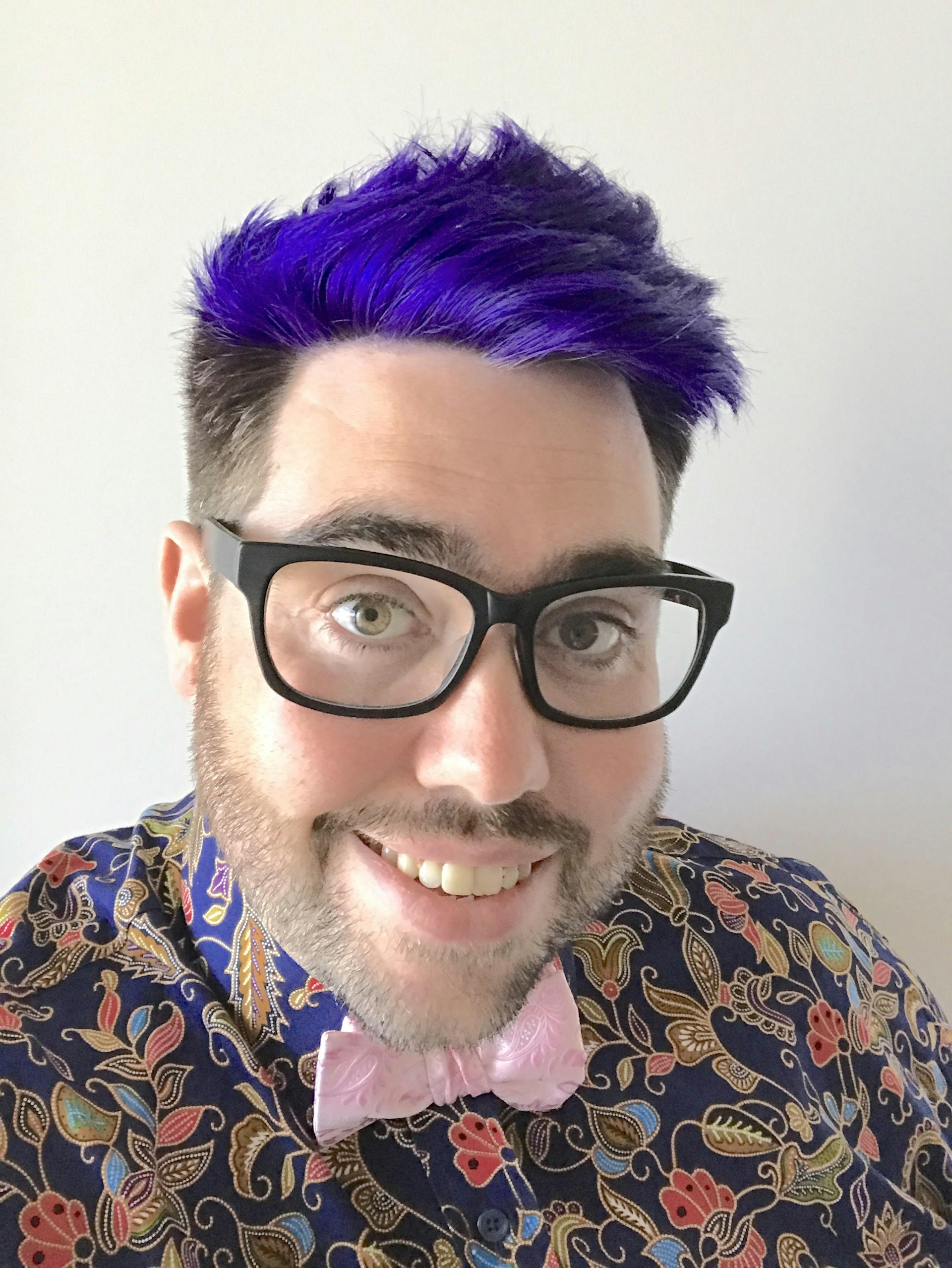 A headshot of a white man with purple hair. He's wearing a patterned shirt, pink bowtie and dark-rimmed glasses, and is smiling at the camera