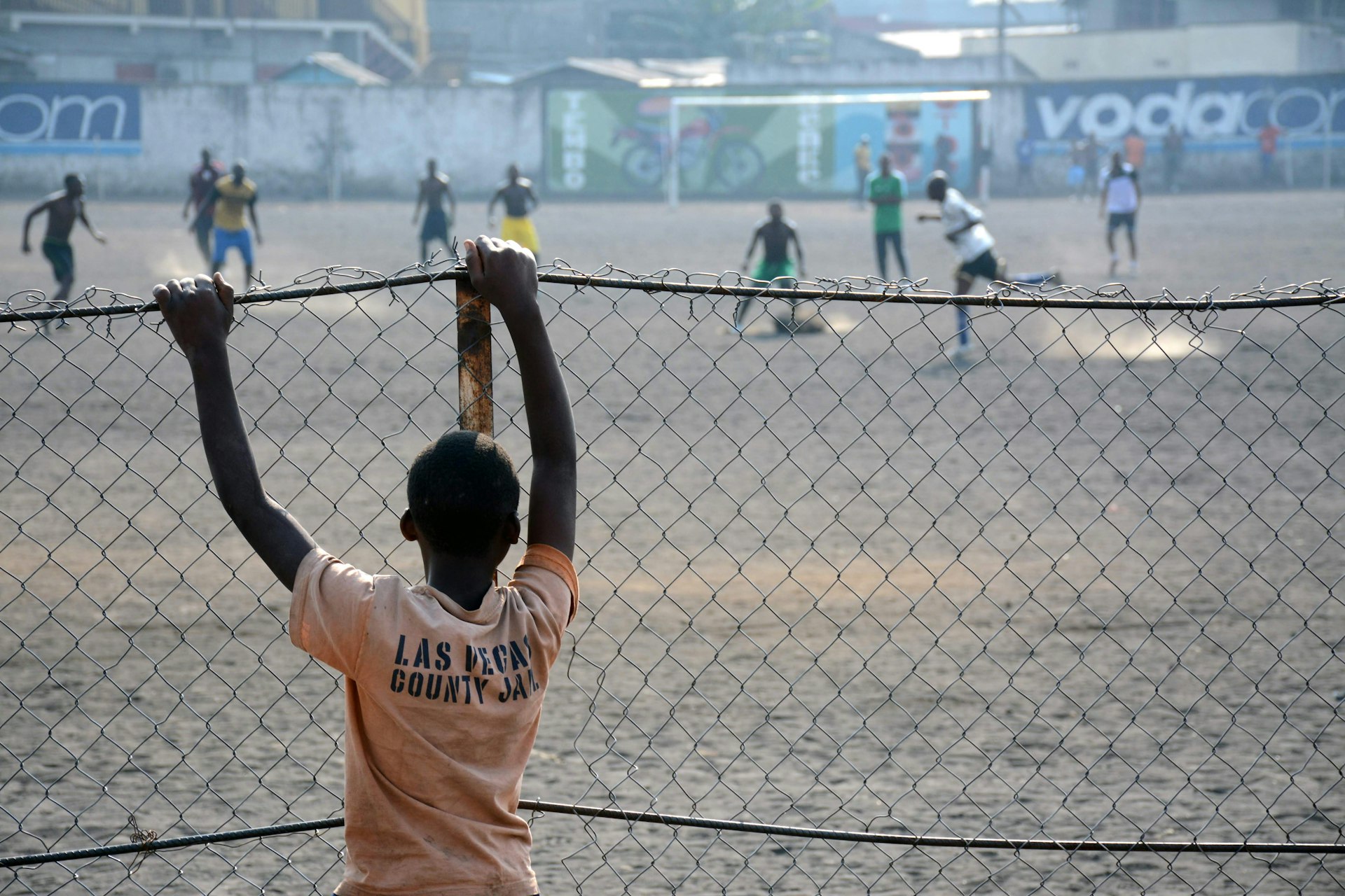 A young boy wearing a faded orange t-shirt watches older men play soccer on a dirt field through a chain-linked fence.