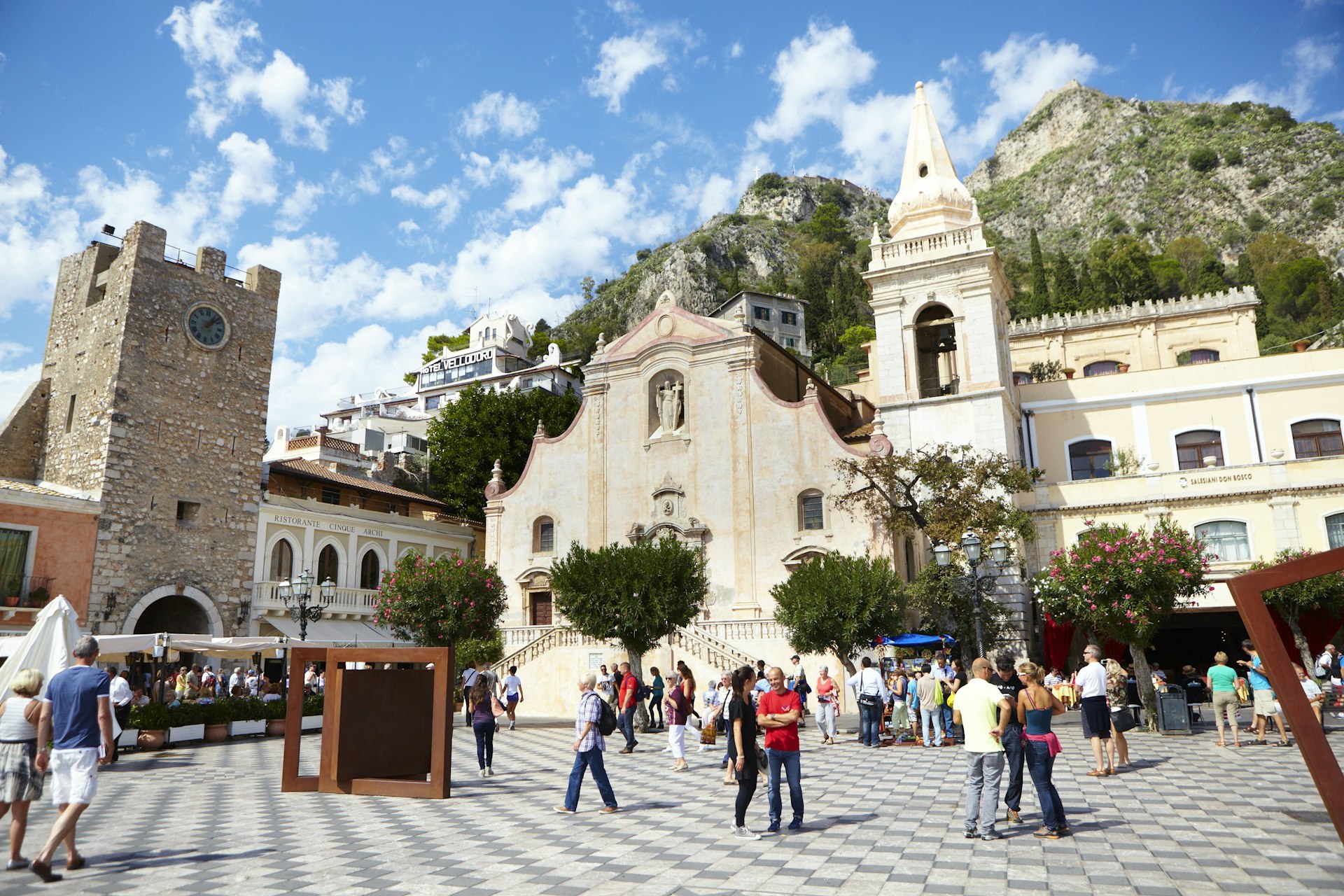 An Italian piazza dotted with people and an ornate church with a mountain in the background