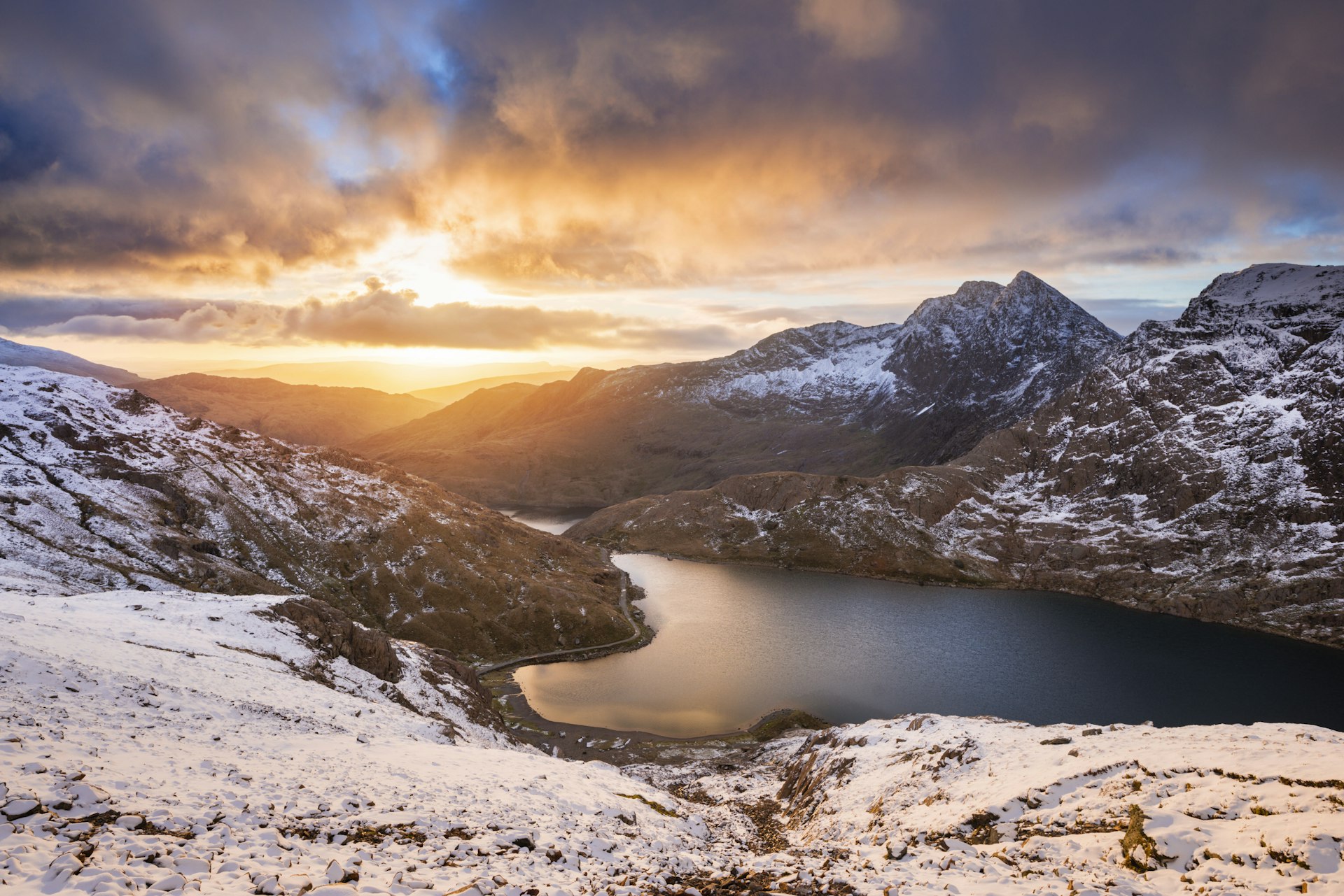 The sun rises over a lake in a snowy mountain landscape, creating orange streaks that contrast with the grey clouds