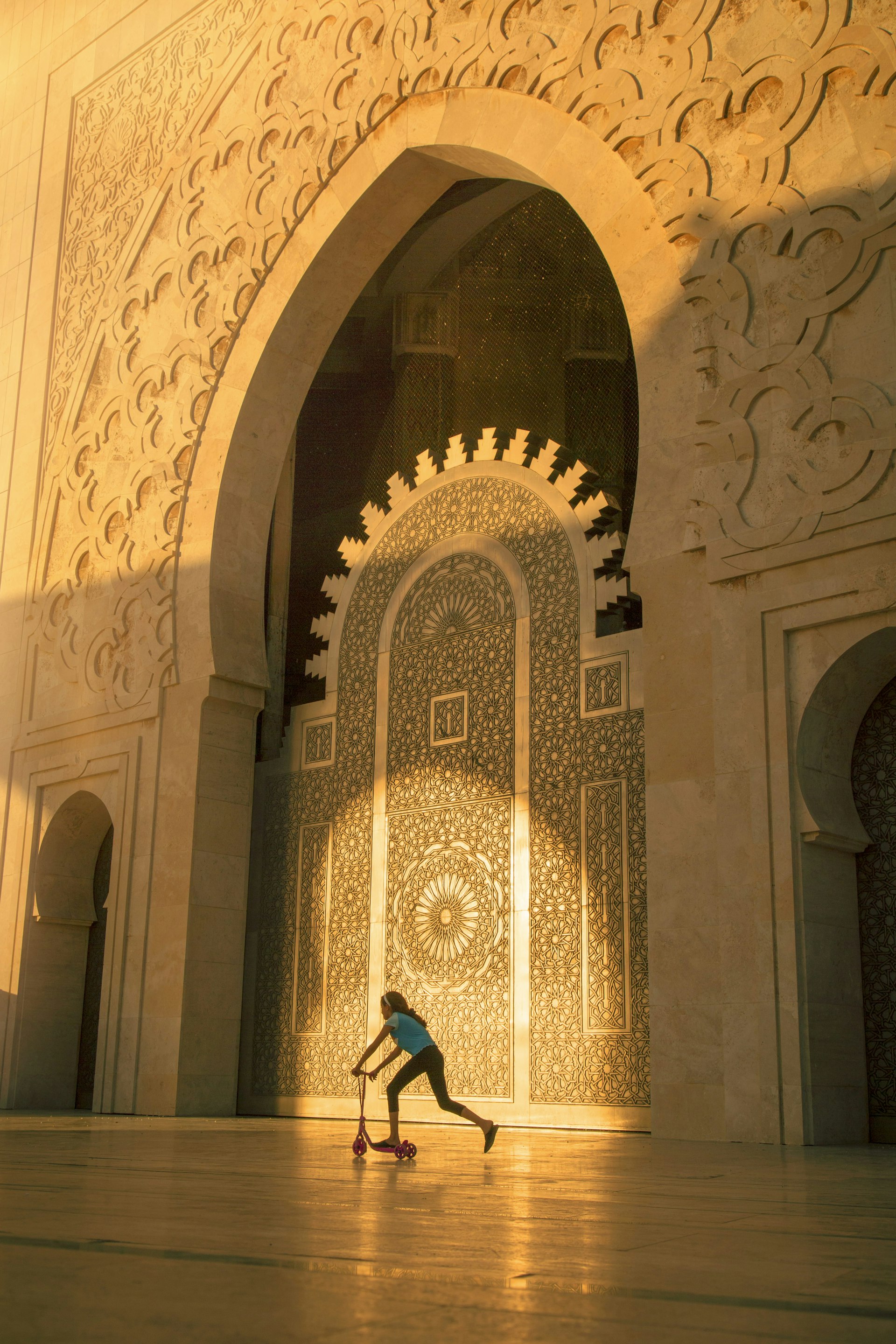 A mosque doorway bathed in golden light, as a young girls zips by on a scooter