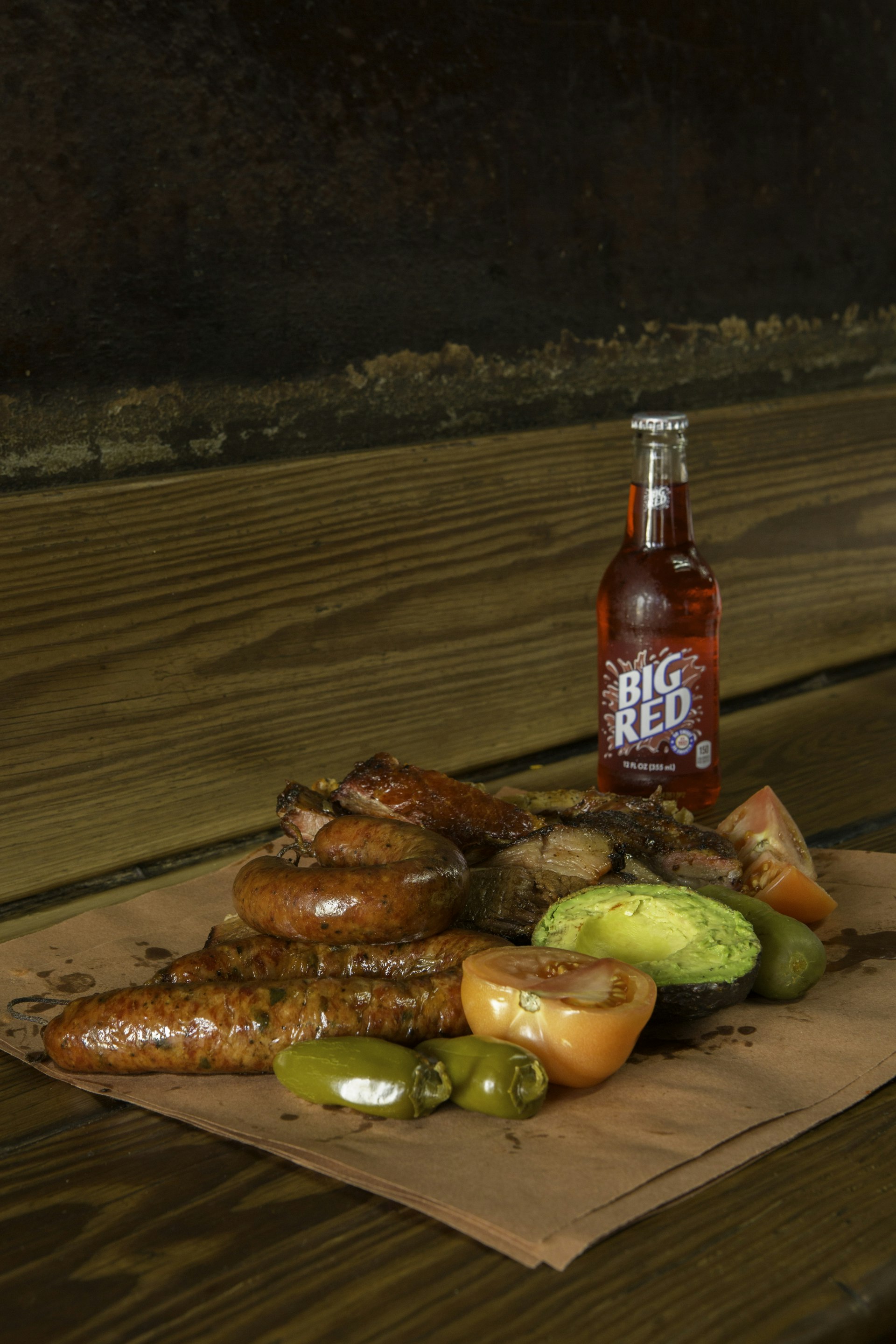 A piece of butcher paper piled with a collection of barbecue meats with tomatoes and peppers. There is a bottle of Big Red next to the food. 