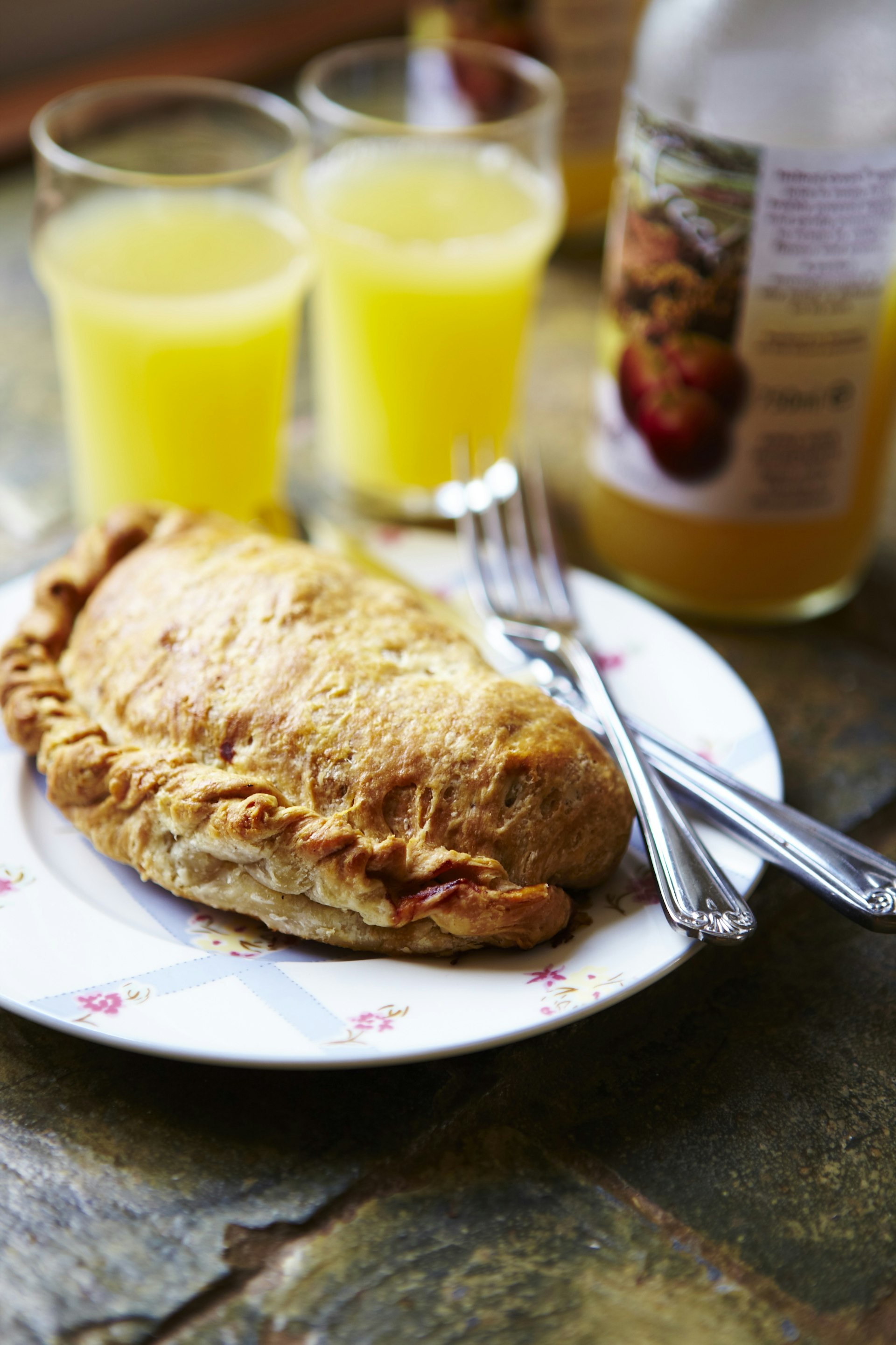 A traditional Cornish pasty on a plate with two glasses of orange juice beside it. The pasty is a baked pastry and filled with potato, meat and vegetables.