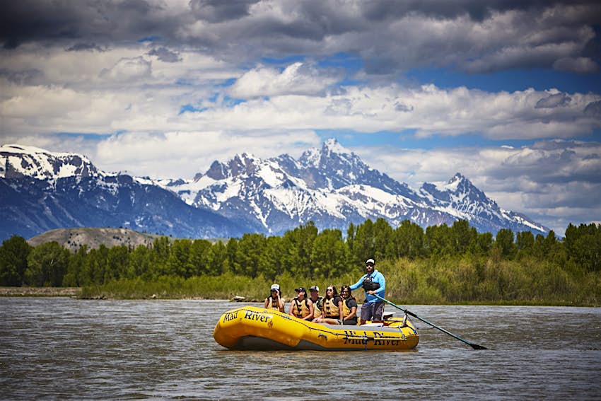 A guide paddles a raft with five passengers on the Snake River, with the Grand Tetons in the distance