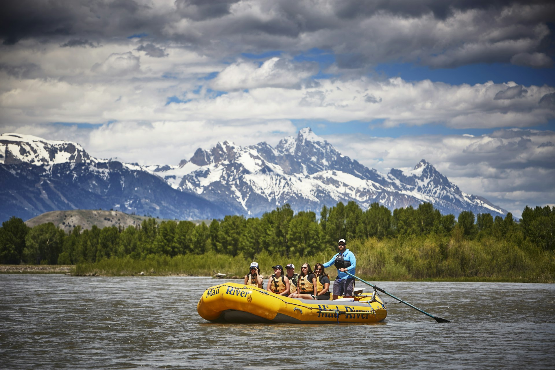 A guide paddles a raft with five passengers down the Snake River, with the Grand Tetons in the distance