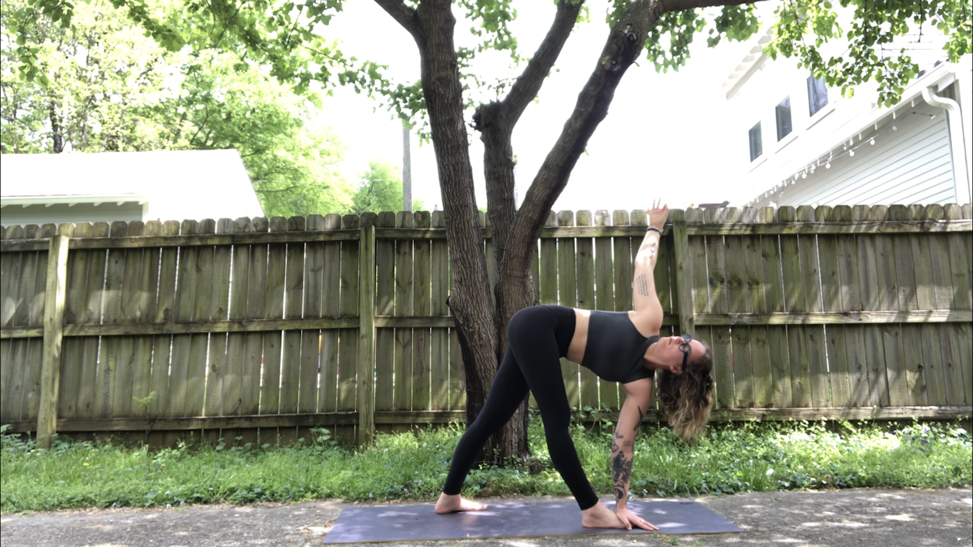 A young woman in black yoga attire demonstrates Parivritta Trikonasna, revolved triangle posture, on a grassy lawn by a worn wooden fence