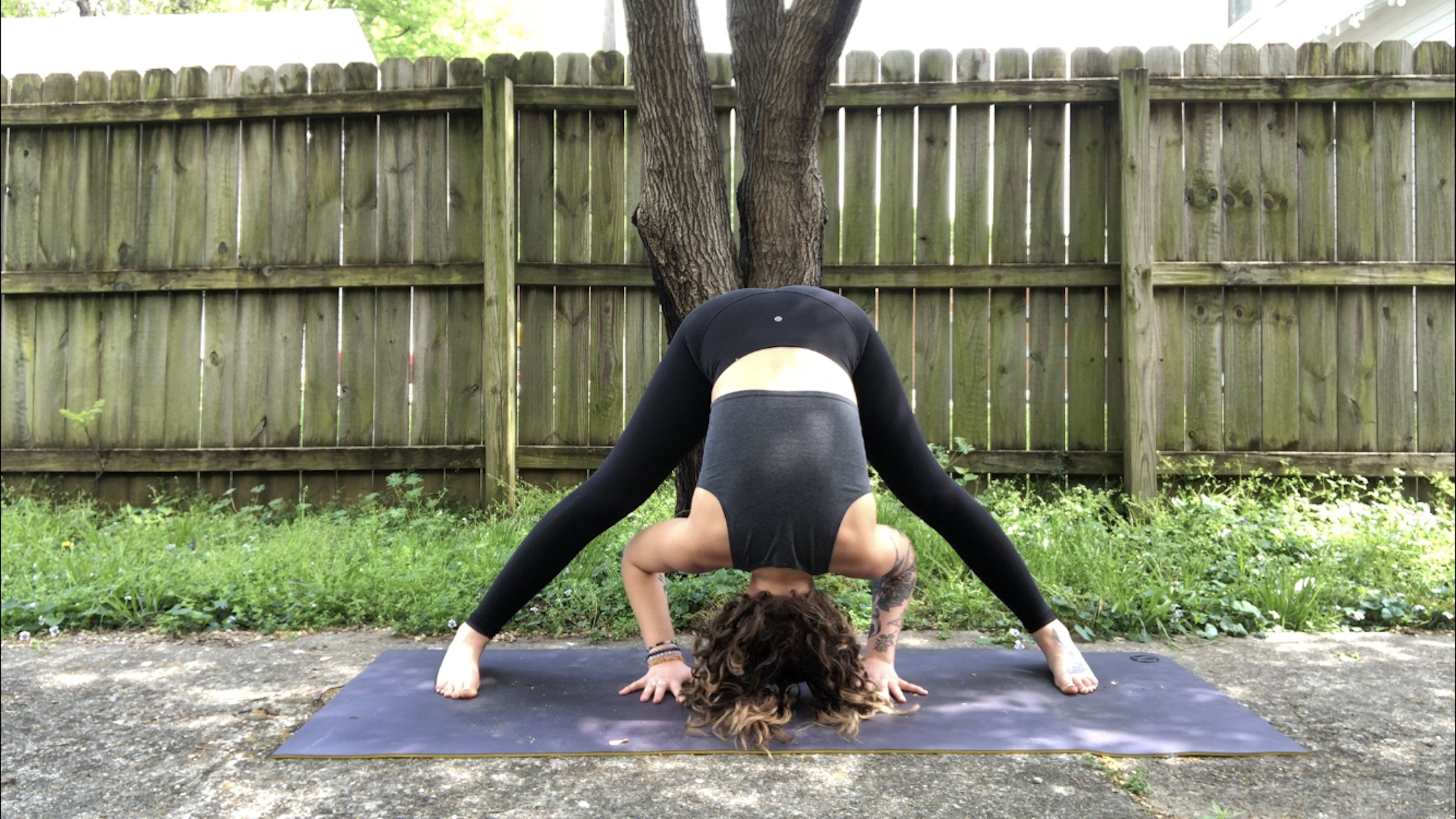 A young woman in black yoga attire demonstrates Prasarita Padottanasana, wide legged forward fold, on a grassy lawn by a worn wooden fence