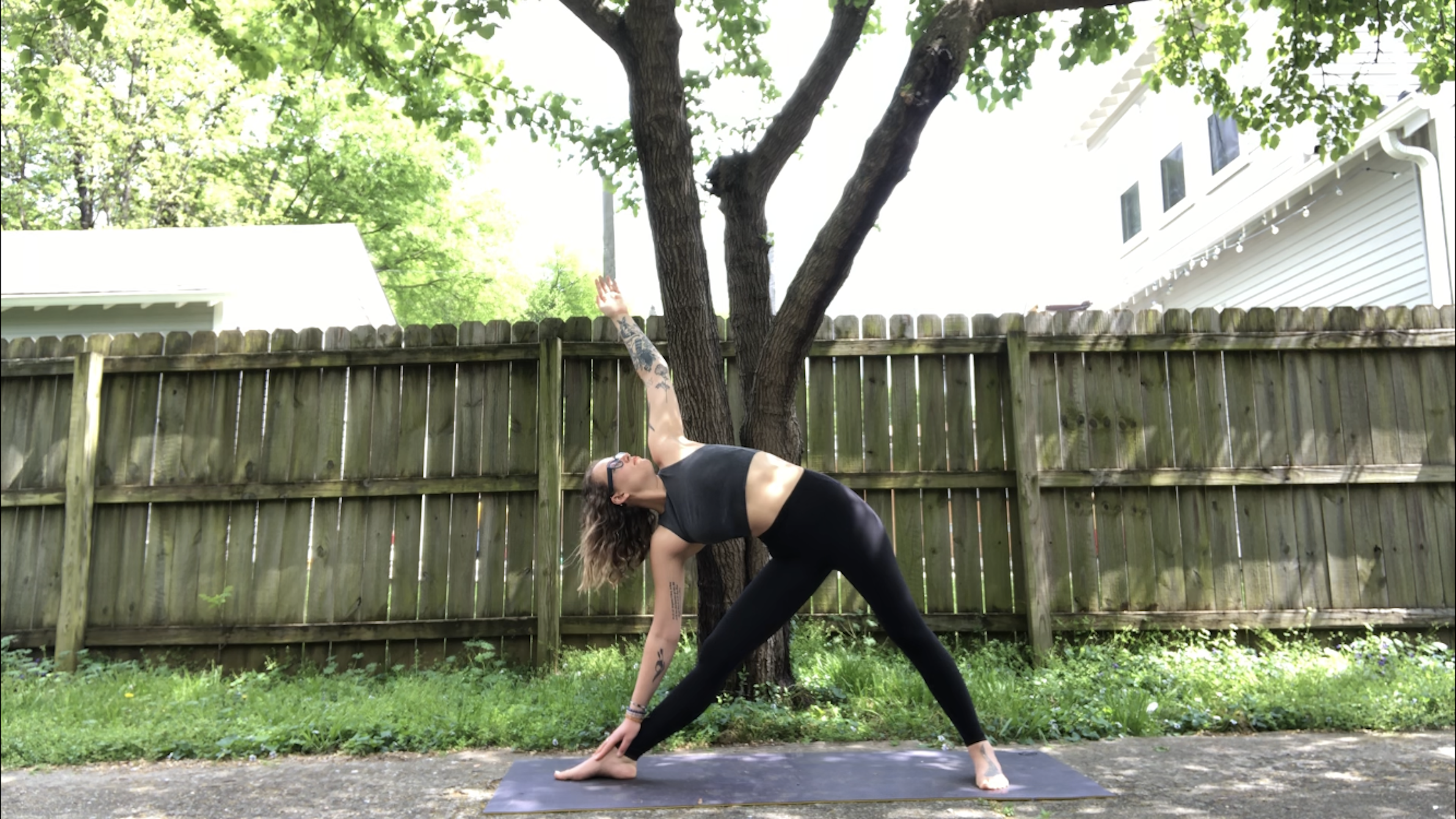 A young woman in black yoga attire demonstrates Utthita Trikonasana, extended triangle yoga pose, on a grassy lawn by a worn wooden fence