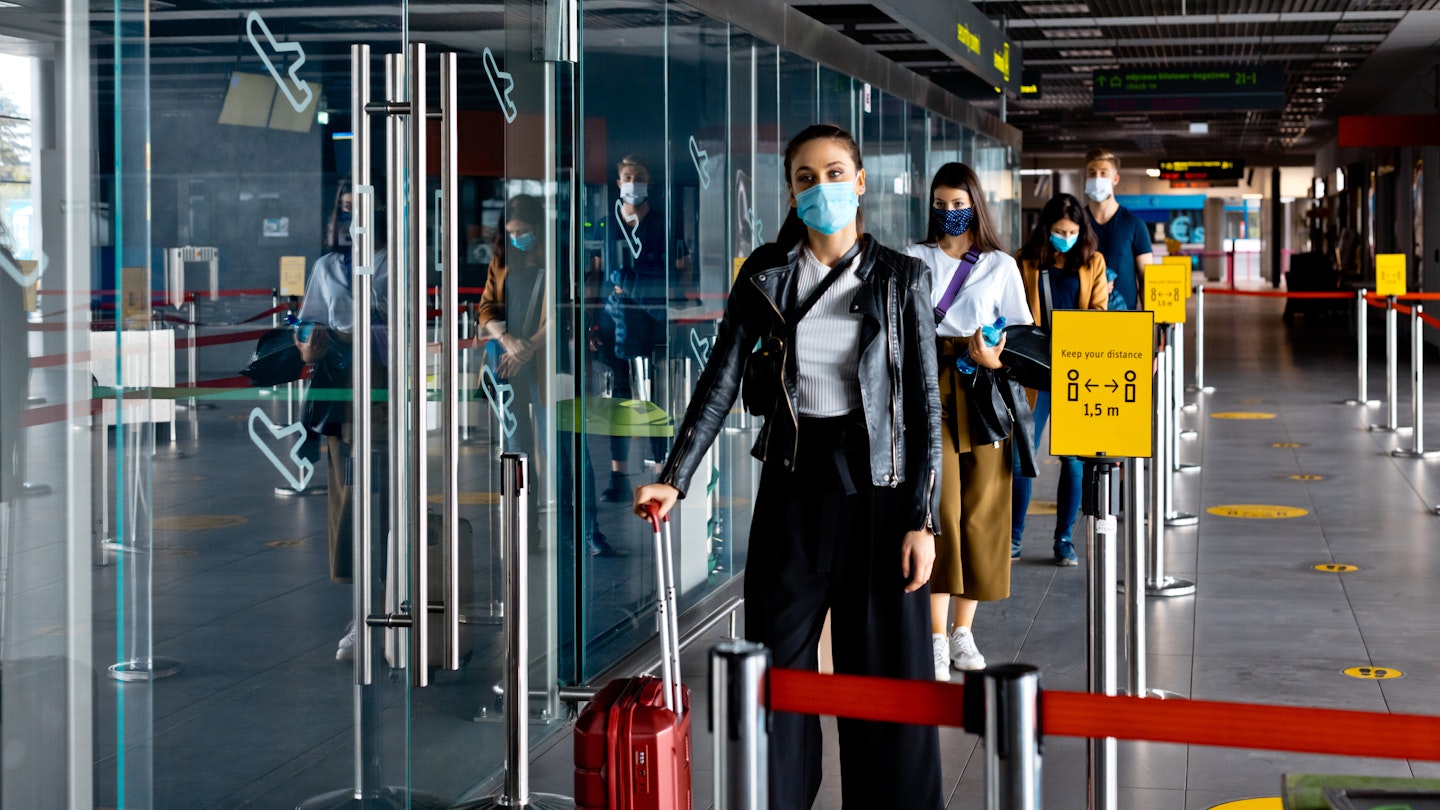 People traveling by airplane during COVID 19, wearing N95 face masks, carrying luggage and waiting in line at airport terminal, keeping distance.