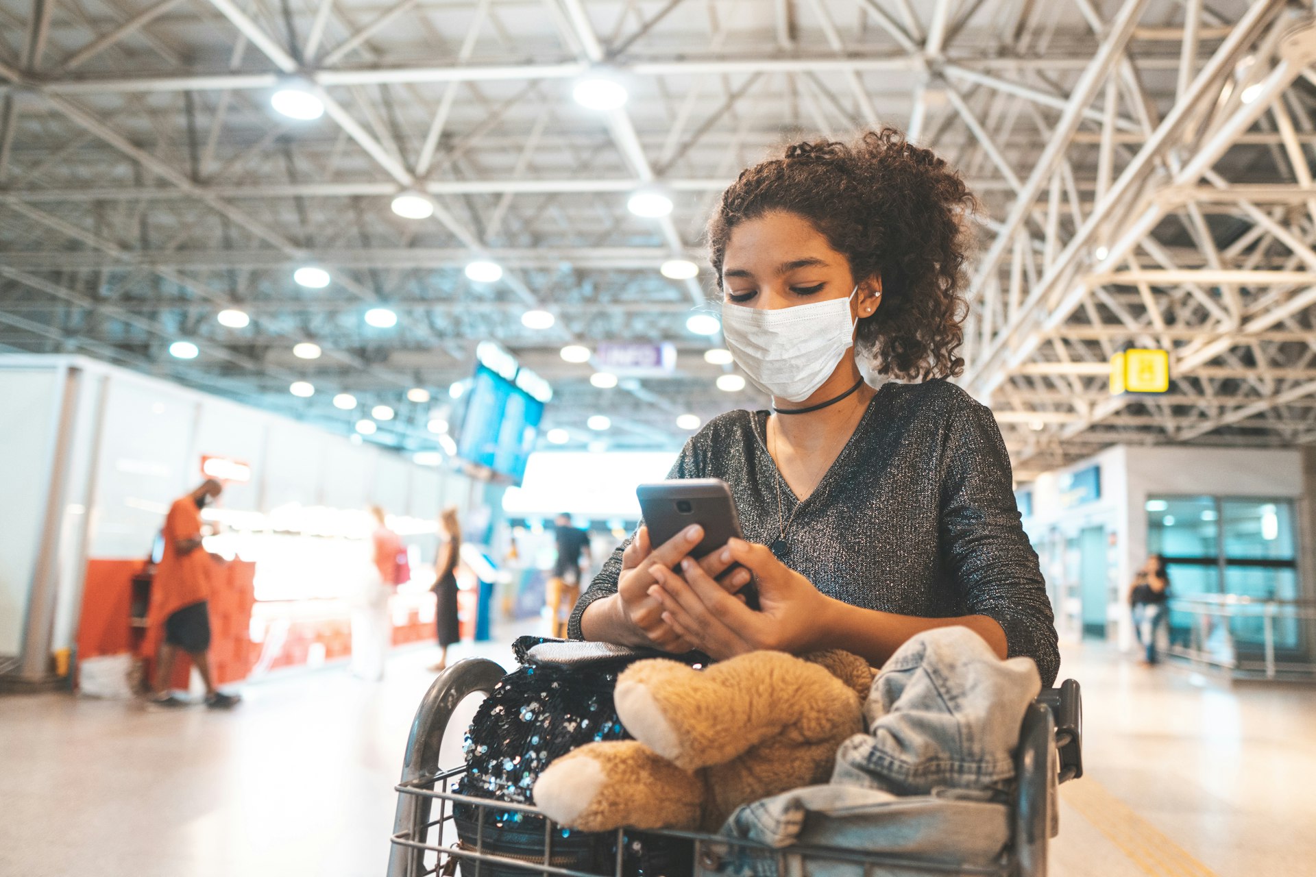 A woman wears a protective mask in an airport. She plays with her phone as she pushes along her luggage trolley