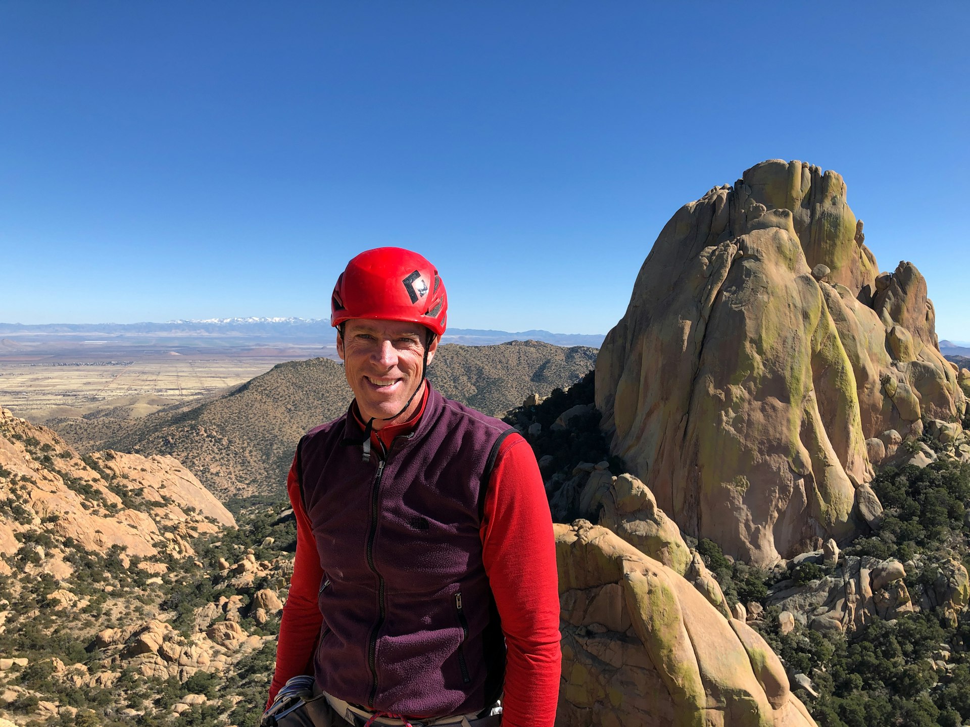 A white man wearing a red helmet smiles at the camera from high altitude with rocky terrain