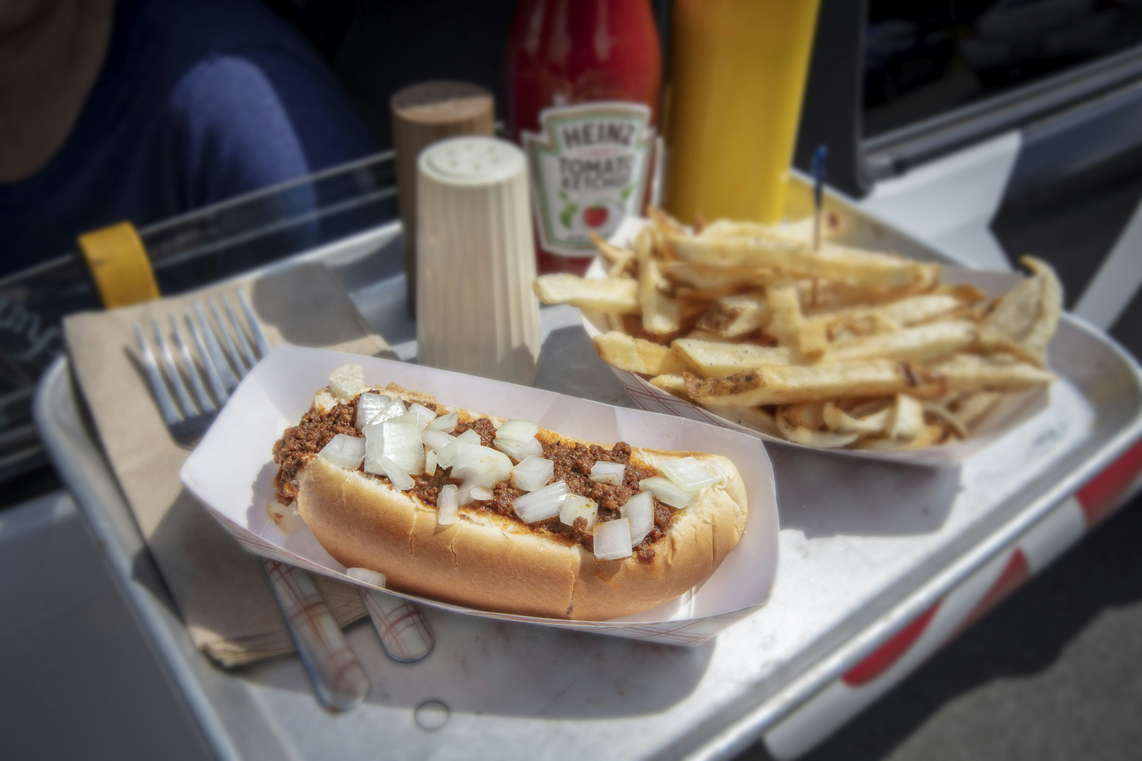 A closeup of a tray with a hotdog topped with meat sauce and onions. There is a side of french fries to the right and in the back is ketchup, mustard with salt and pepper