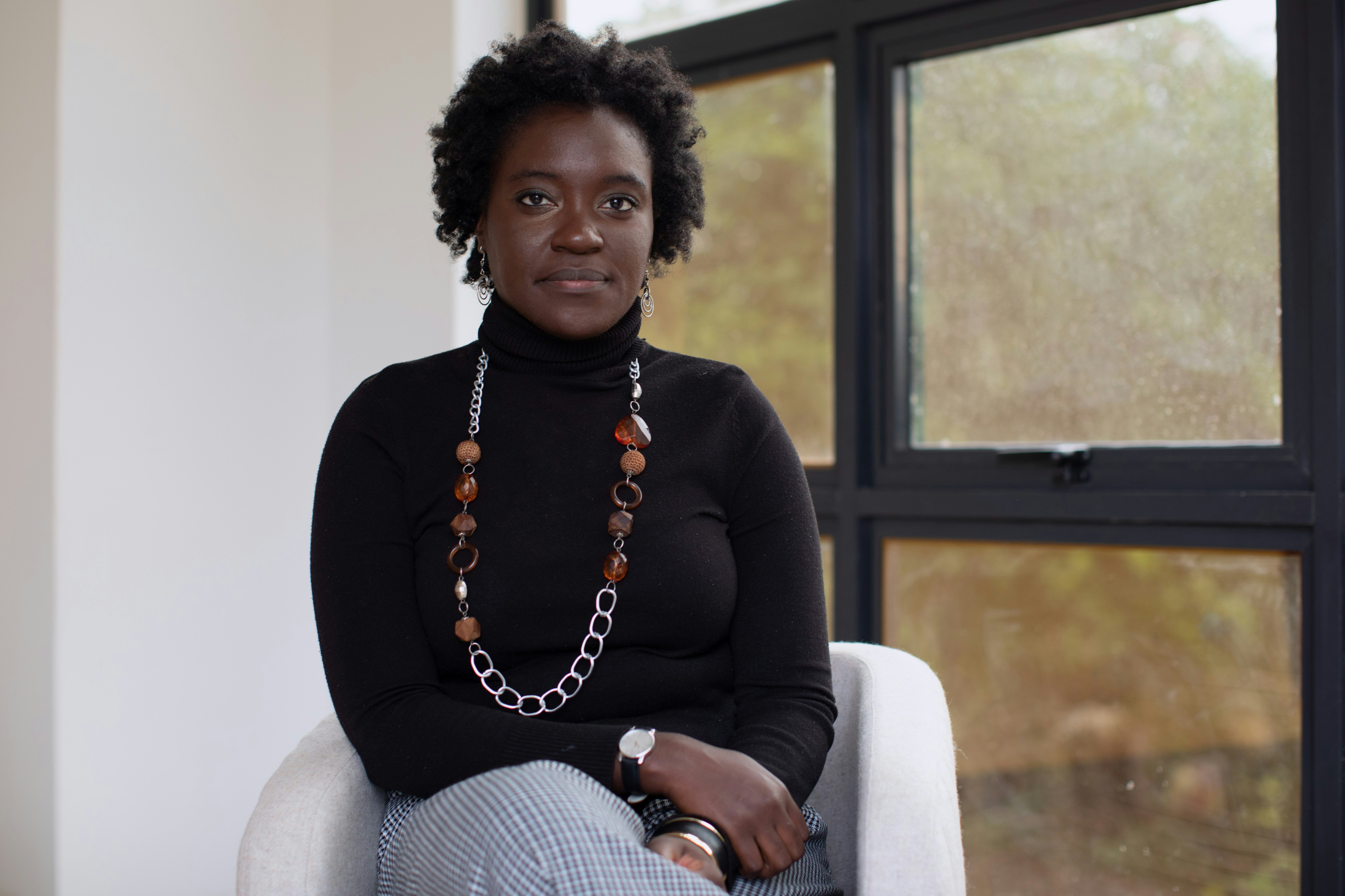 A black woman sits in a chair by a window and looks into the camera