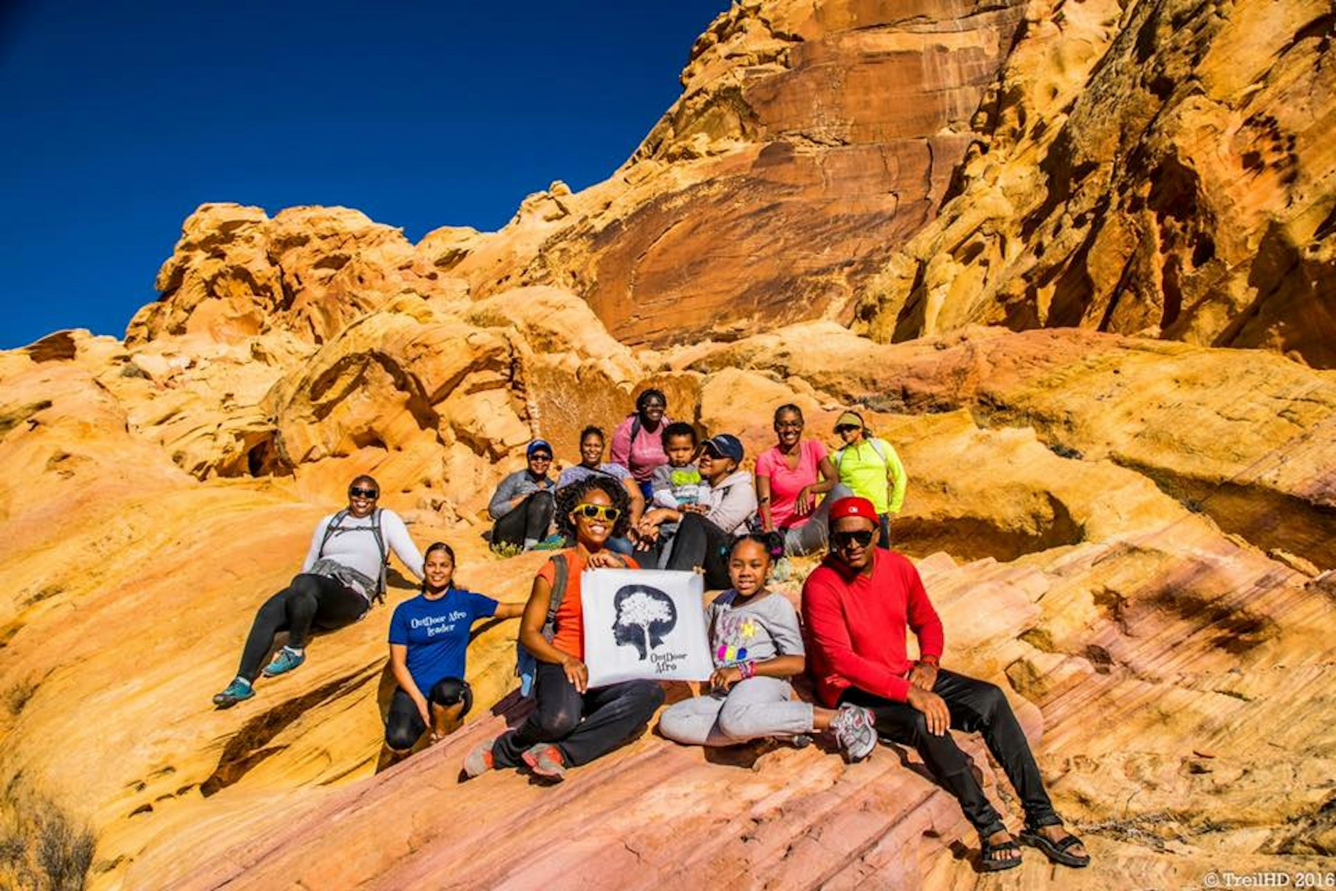 A group of black hikers pose with a small Outdoor Afro banner on the side of sand-colored rocks