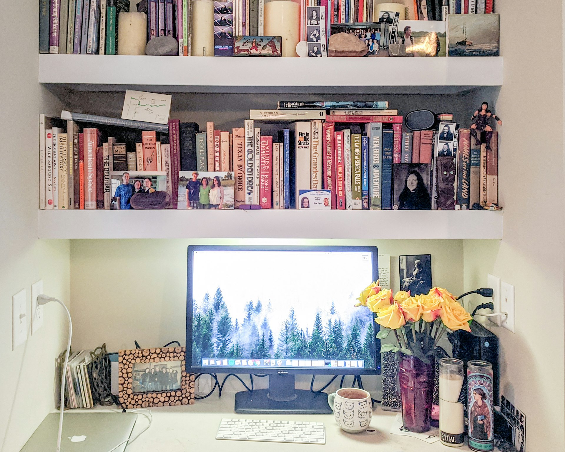 A home office setup that consists of a desk under a bookshelf full of colorful volumes. A vase of yellow roses sits on the desk next to a computer monitor, keyboard, mouse, and laptop, surrounded by framed photos and mementos