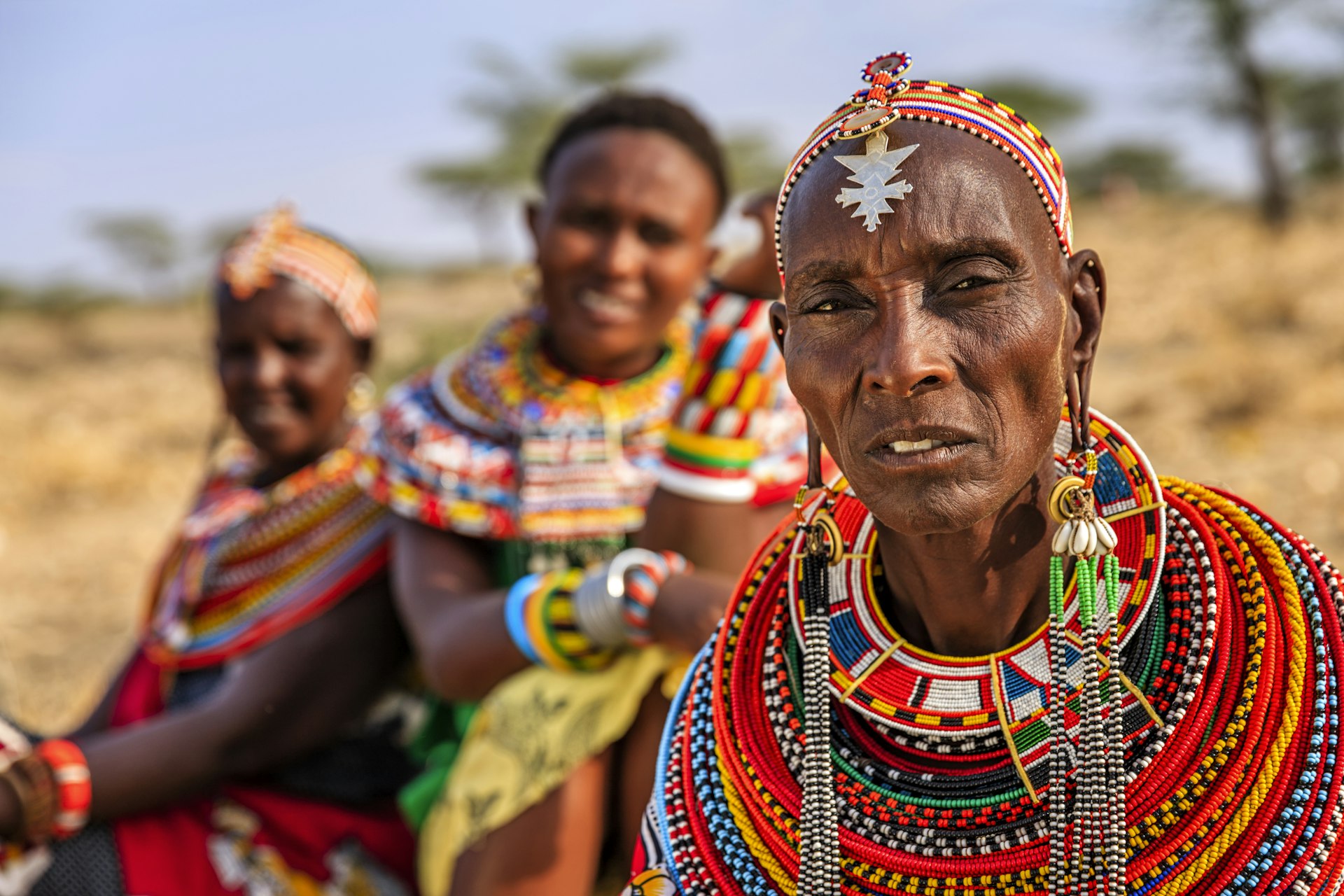 Three women in the Kenya desert look at the camera, one is in focus and two in the background are not. The woman in front wears a headdress with a pendant and is wearing dozens of beaded necklaces.