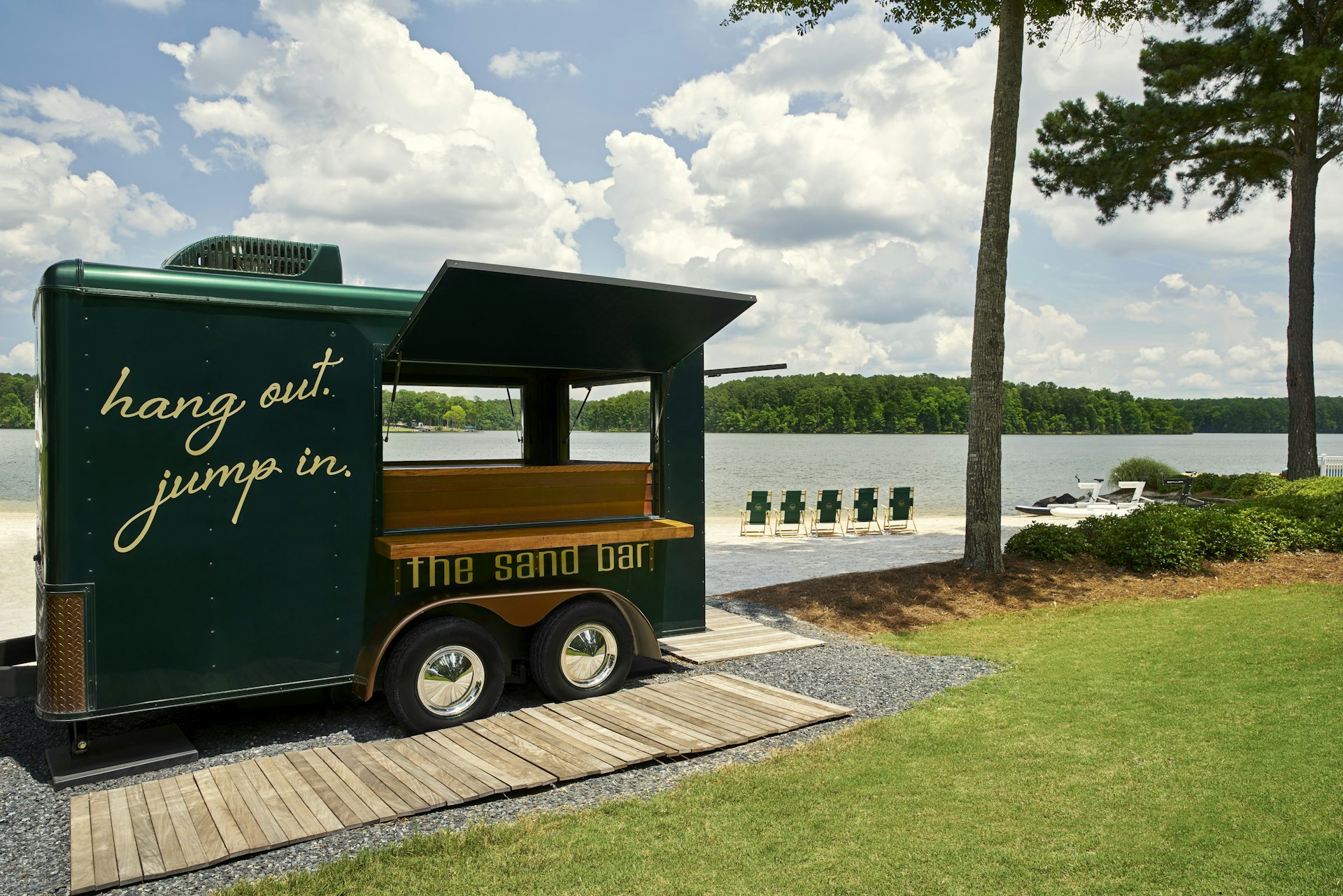 A mobile bar with the words "hang out, jump in" painted on the side is parked by the side of a lake, with several sunloungers nearby