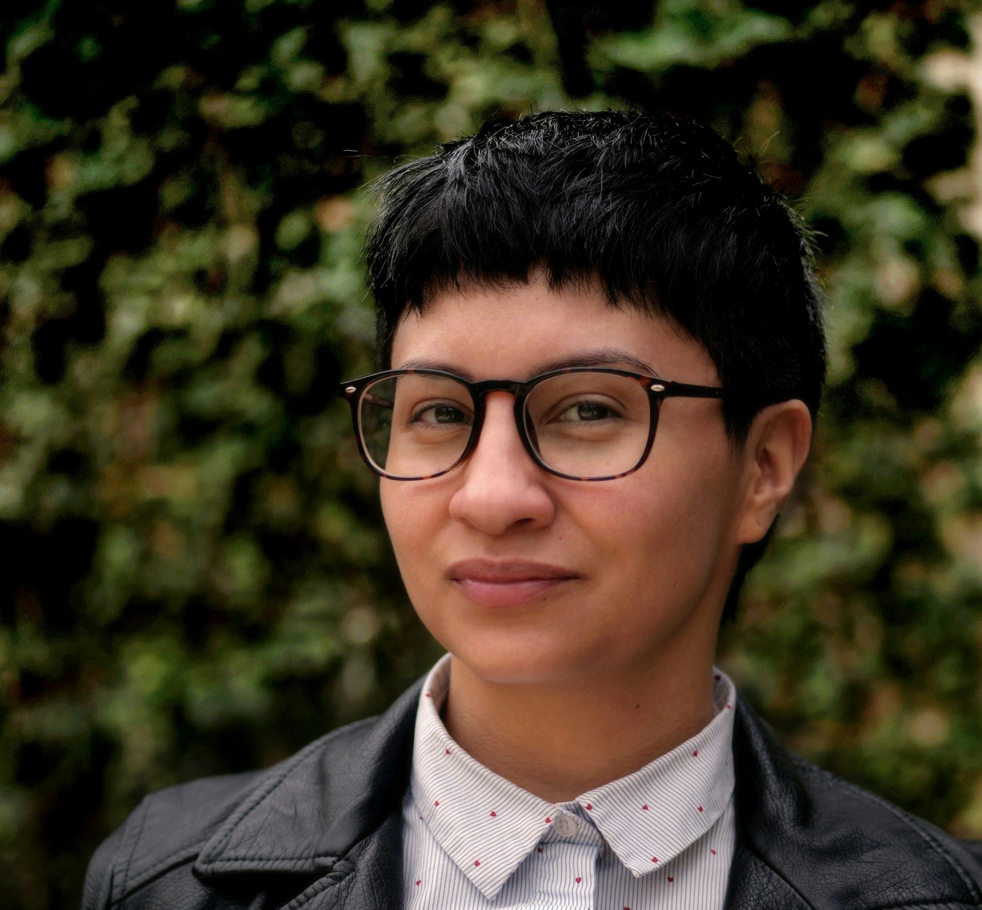 A gender/queer Latinx person of Native and white descent, wearing black-rimmed glasses, smiles for the camera